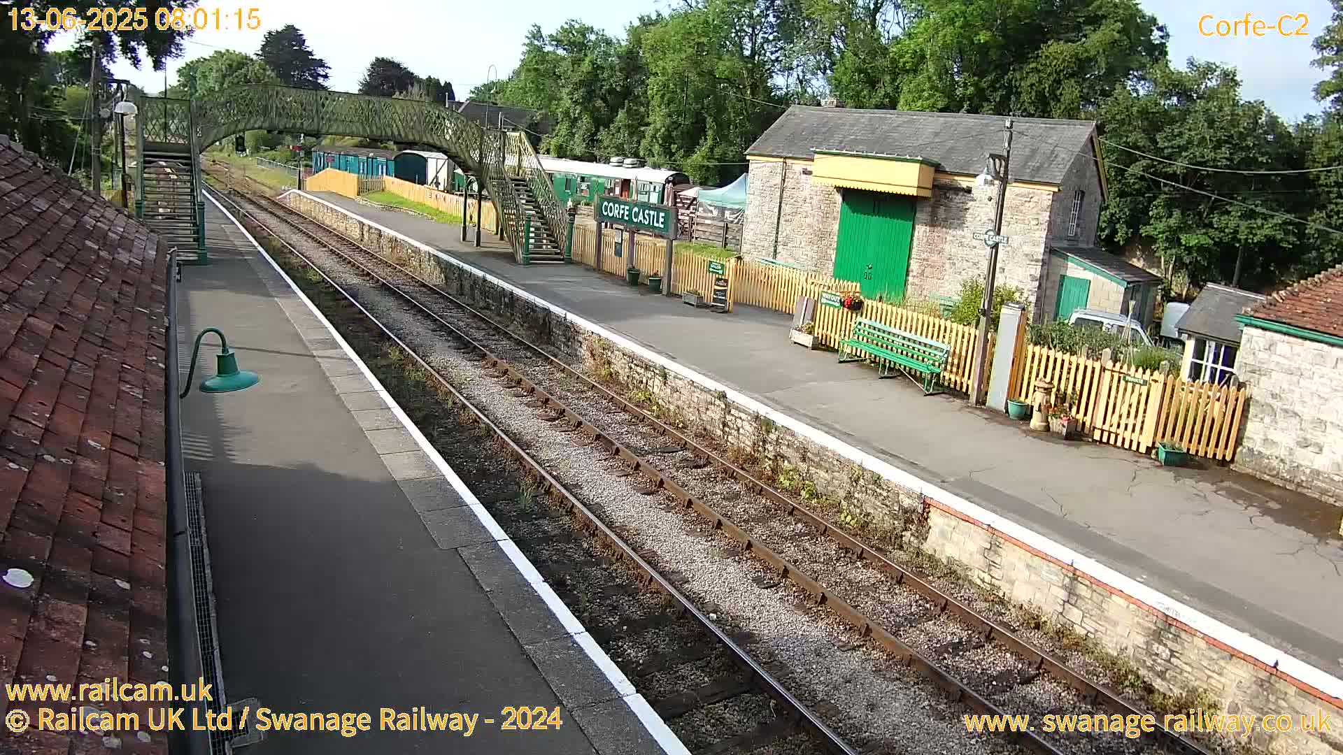 A railway station platform with train tracks, a stone building, and a footbridge under a sunny sky.