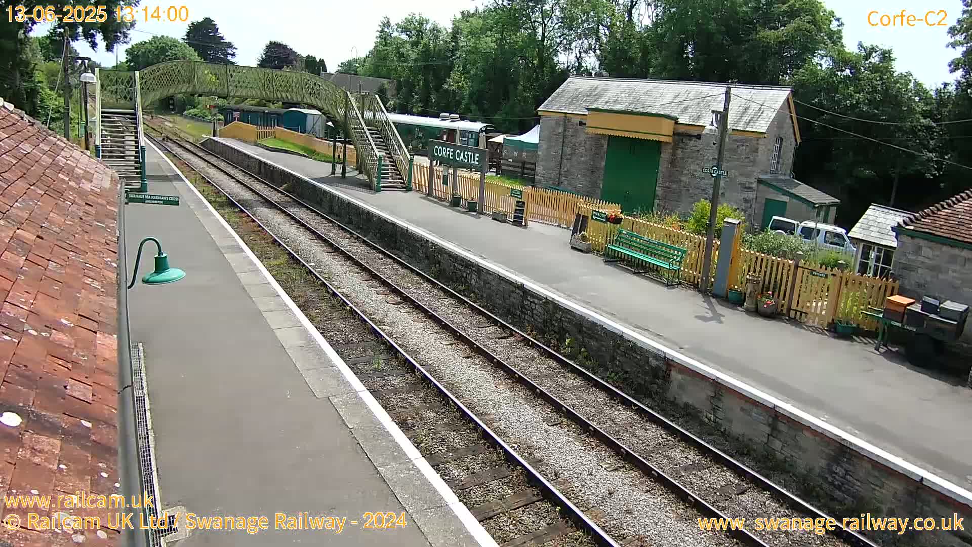 A train station platform with railway tracks, stone buildings, and a footbridge on a sunny day.