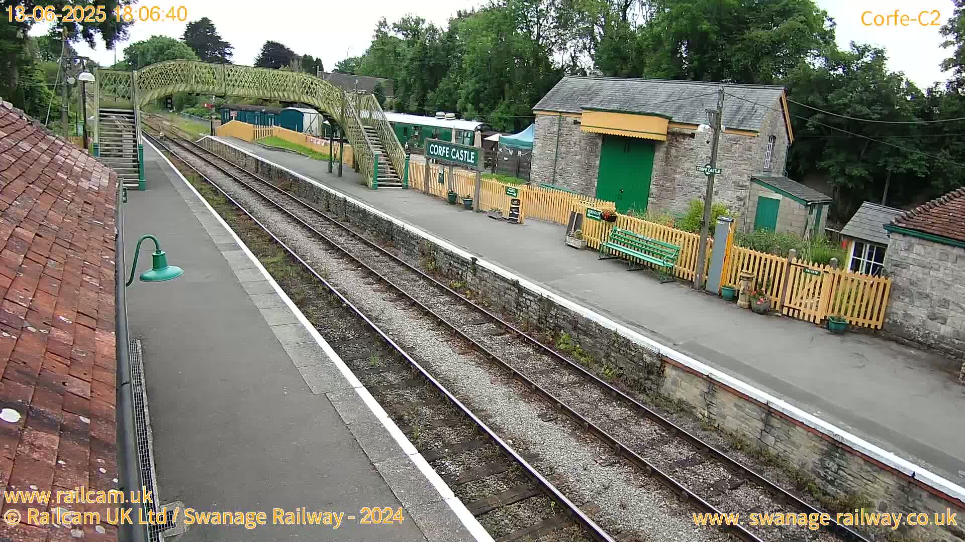 A train station platform with a footbridge, stone buildings, and train tracks under a partly cloudy sky.