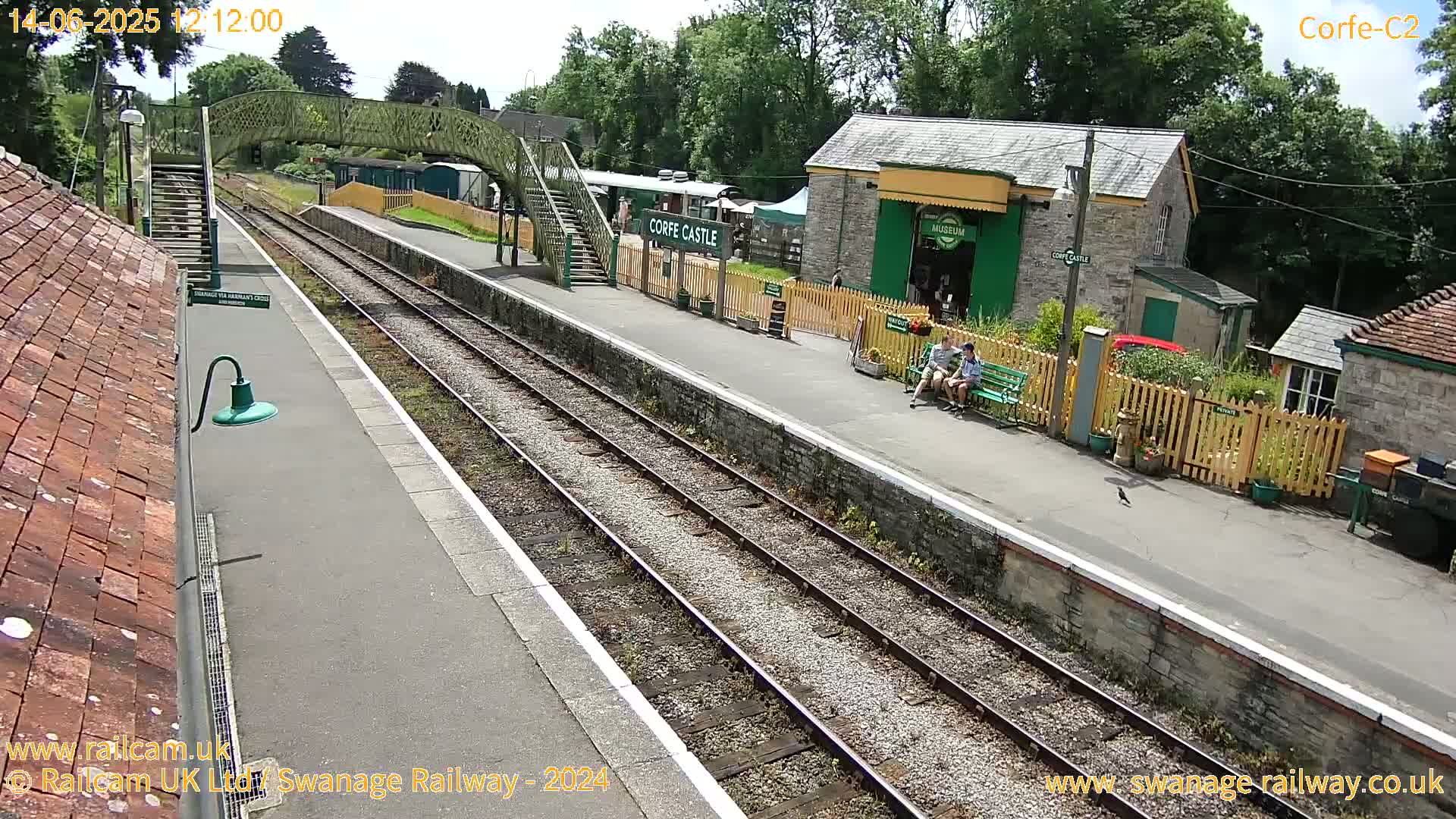 A sunny day at a train station shows two parallel train tracks, a platform with a few people, station buildings, and a footbridge.