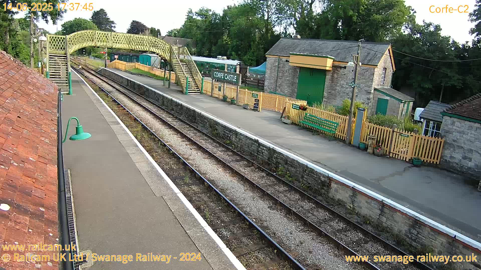 A train station platform with railway tracks, a pedestrian bridge, and stone buildings under a partly sunny sky.