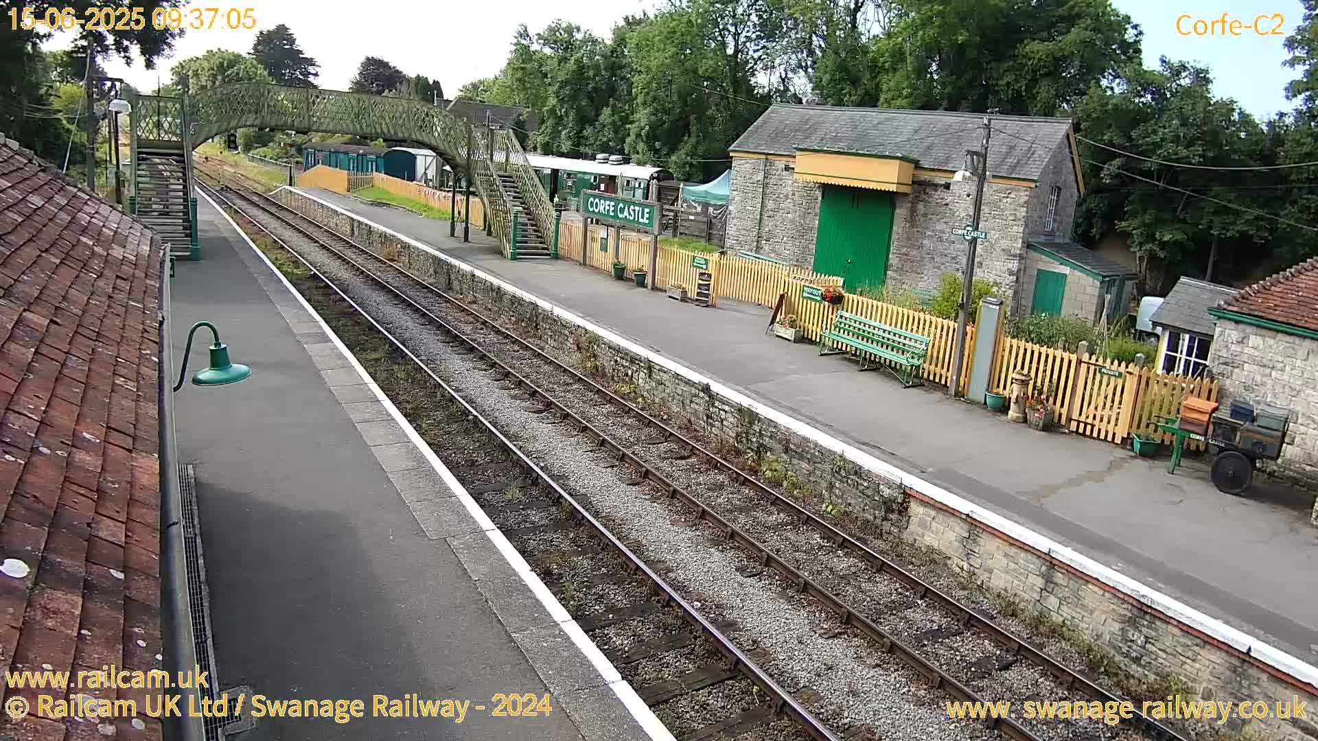 A railway station platform with a pedestrian bridge, stone buildings, and train tracks under a partly sunny sky.