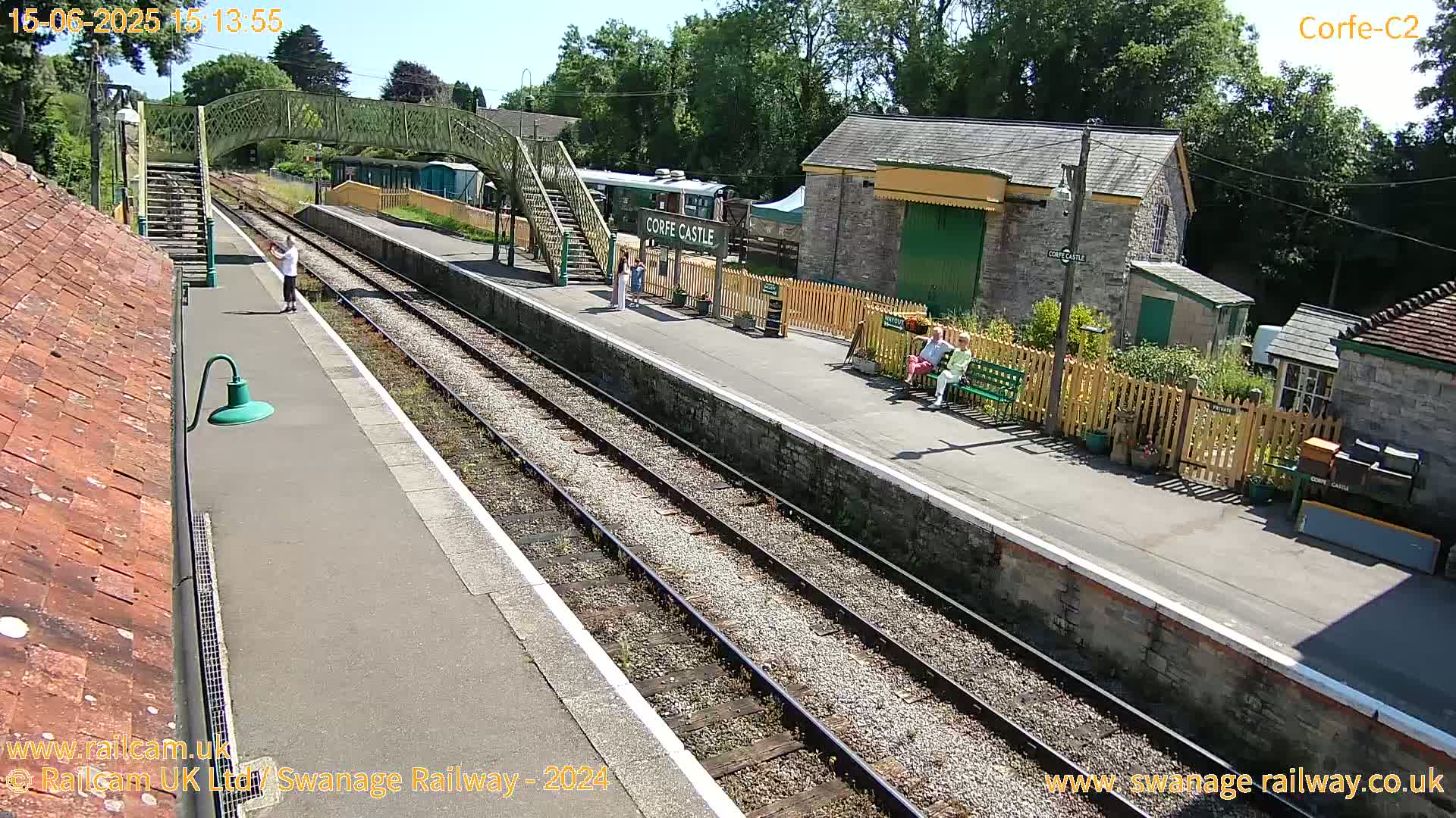 A sunny day at a train station shows a platform with several people, train tracks, a footbridge, and station buildings.