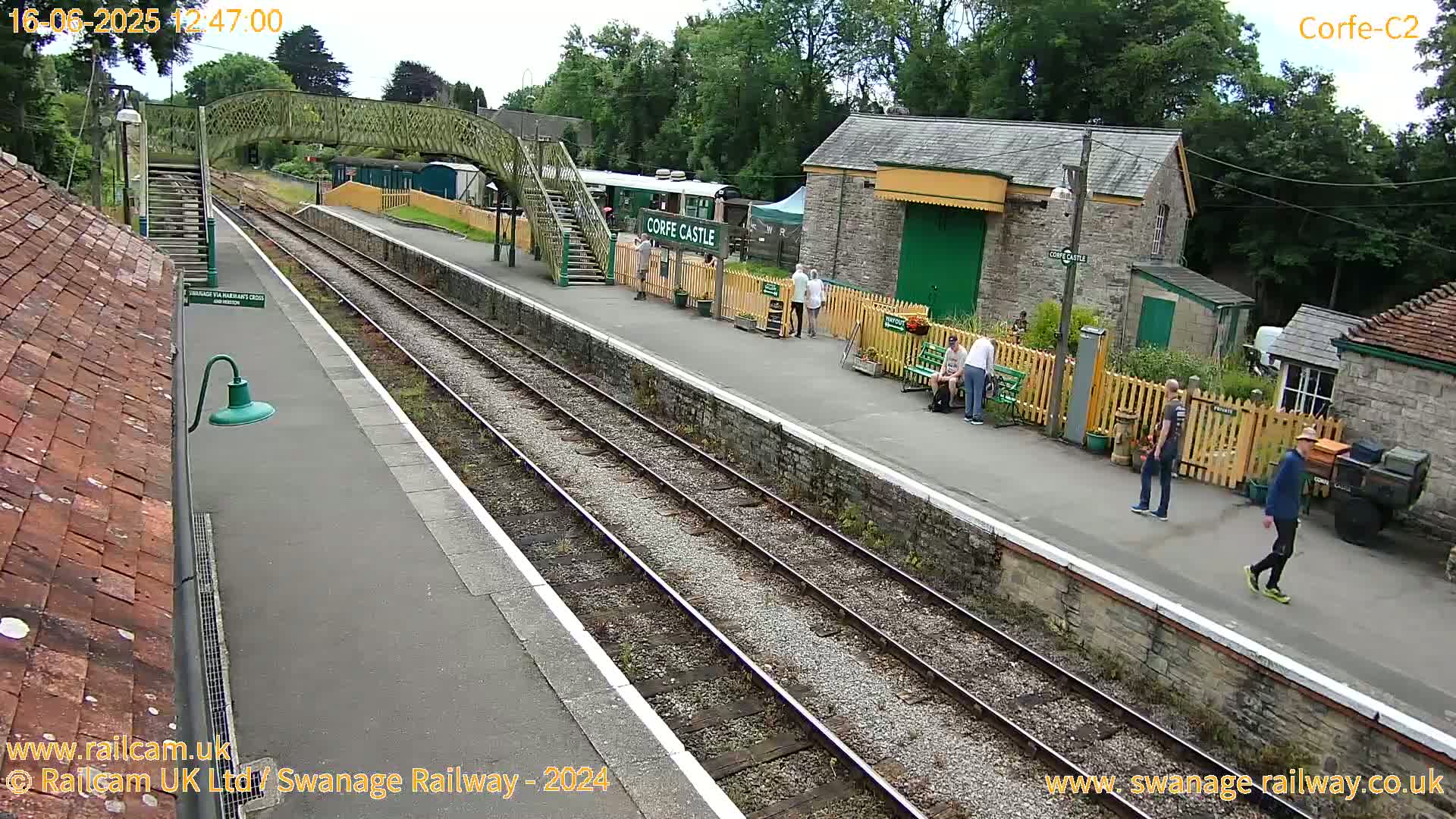 A railway station platform on a partly sunny day shows several people standing and sitting near railway tracks and buildings.