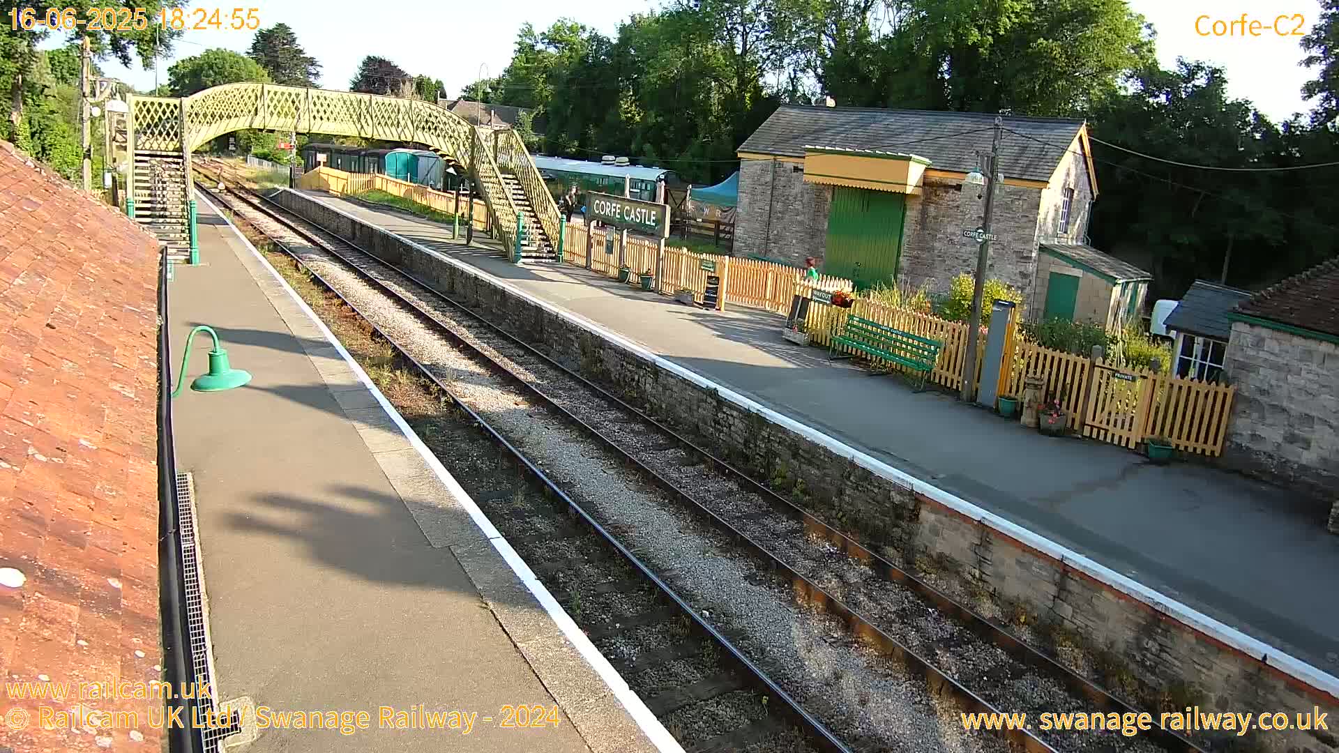 A sunny view of a train station with two sets of tracks, a pedestrian bridge, and several stone buildings.