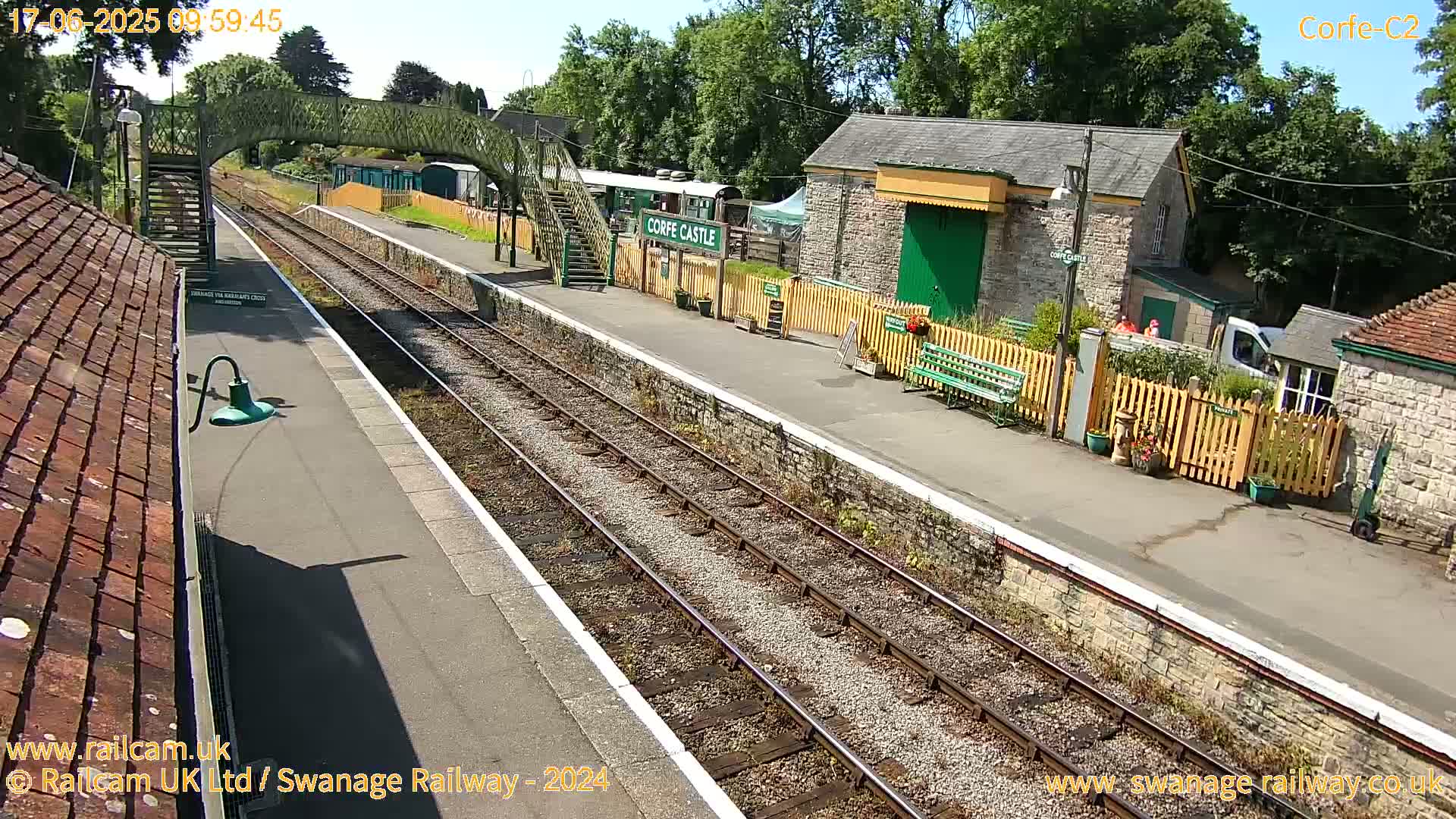 A sunny view of a train station platform with railway tracks, a stone building, and a footbridge.
