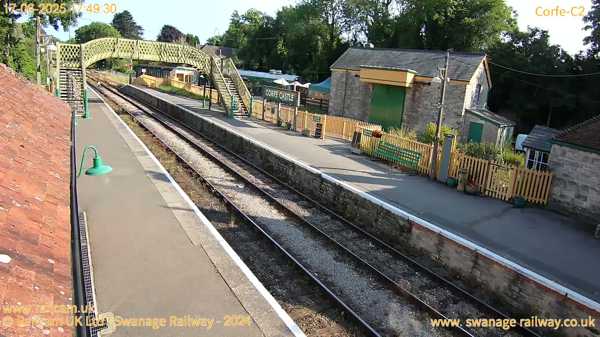 A train station platform with two train tracks, a pedestrian bridge, and several small buildings is shown on a sunny day.
