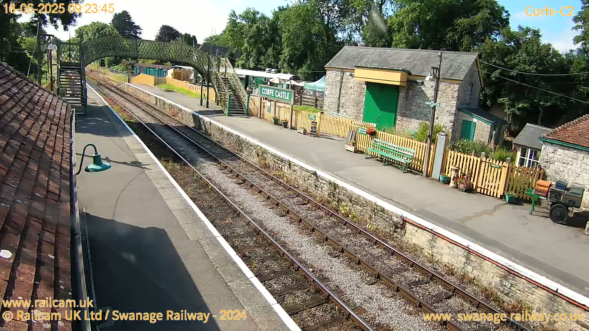 A sunny day at a train station with a footbridge, stone buildings, and train tracks.