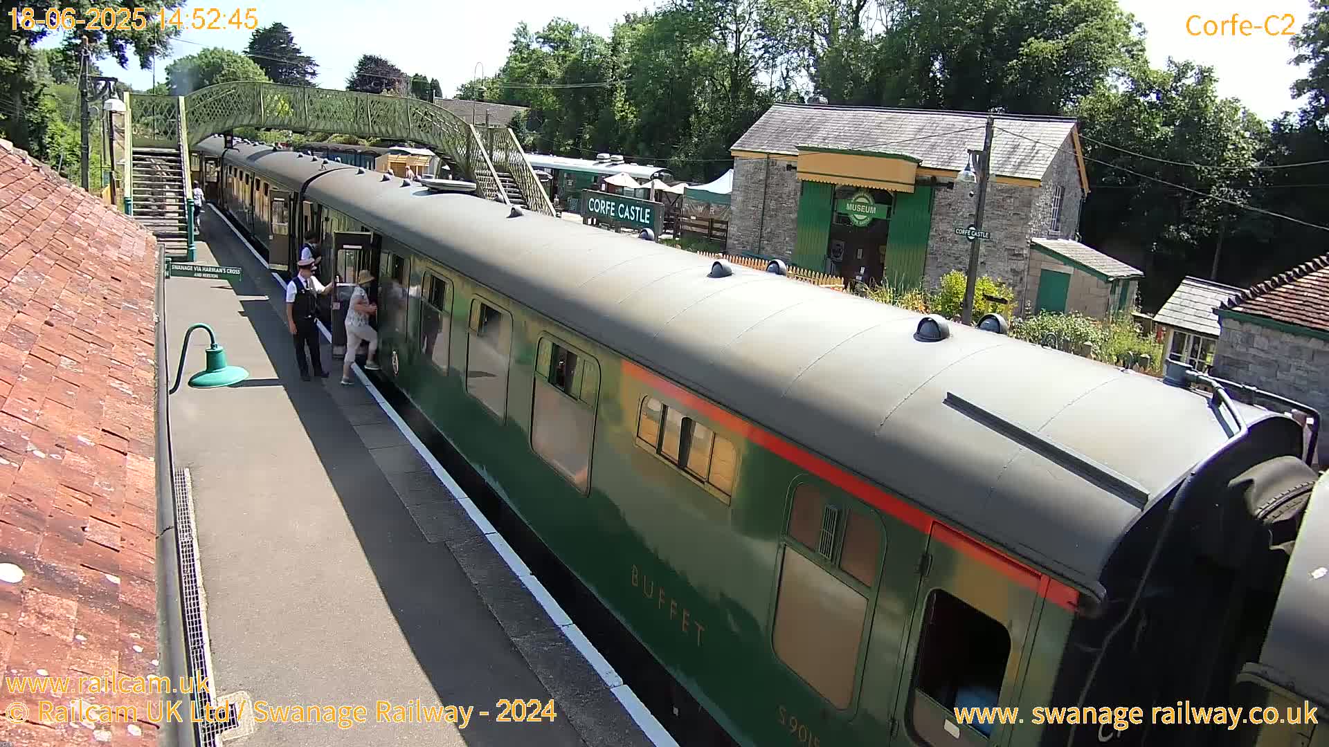 A green passenger train is stopped at a station on a sunny day, with people getting on and off.