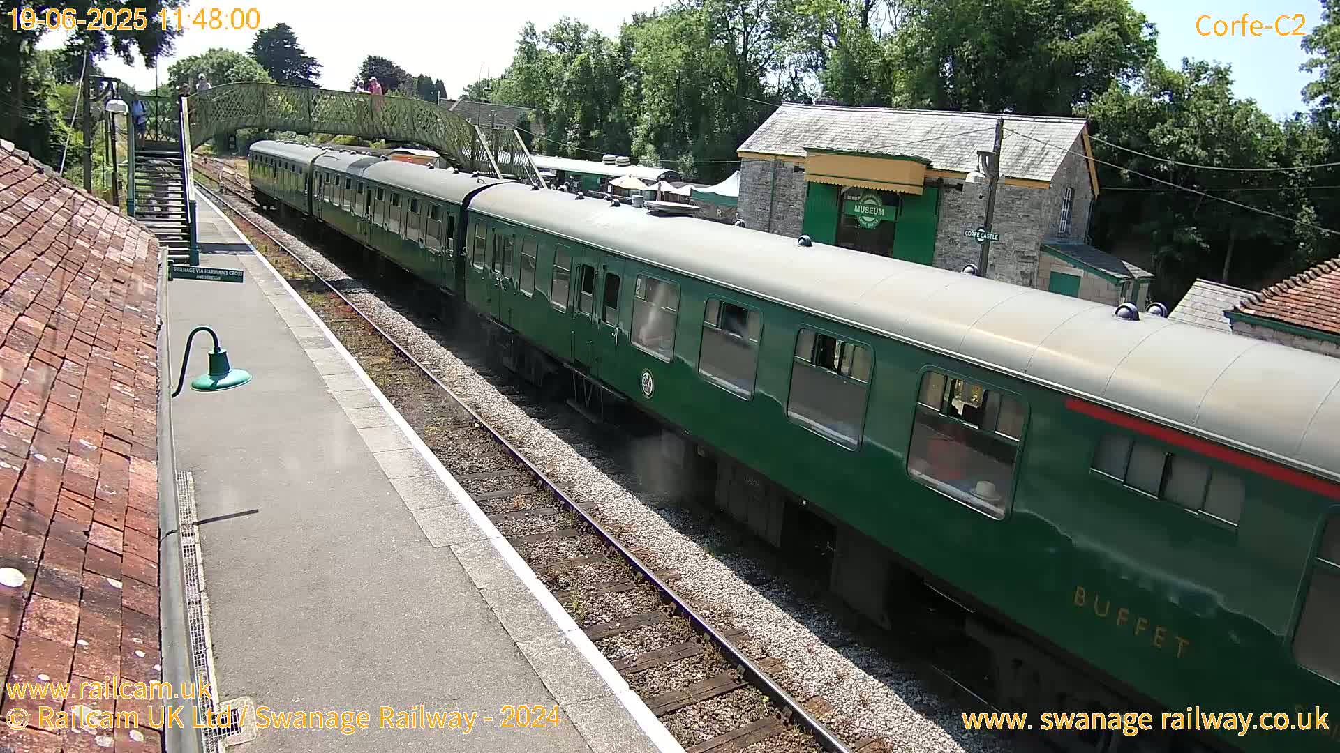 A green passenger train is departing a station on a sunny day.
