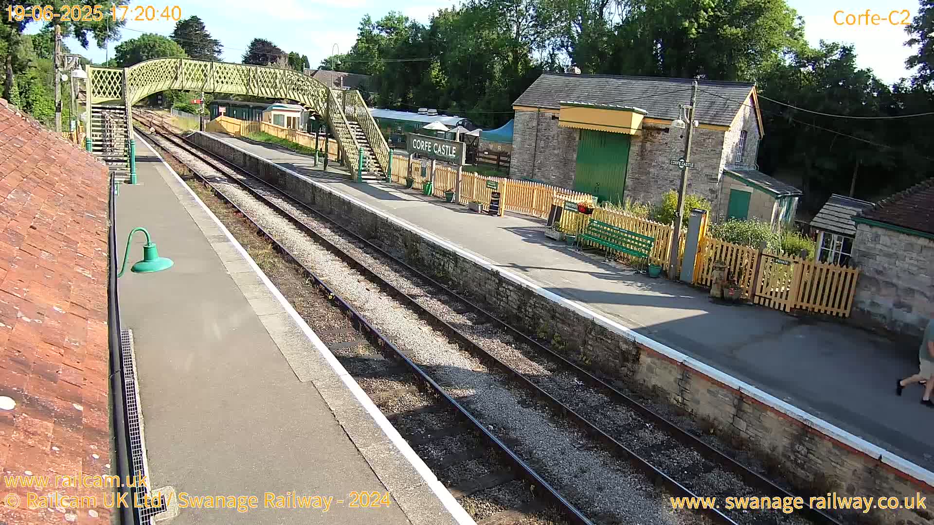 A sunny train station with a footbridge, railway tracks, platform, and stone buildings.