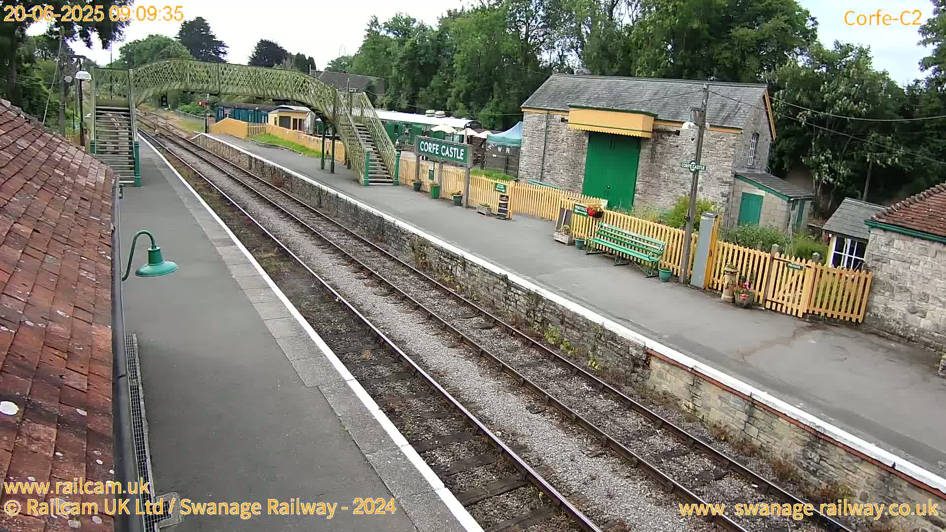 A train station platform with multiple train tracks, stone buildings, a footbridge, and a partially overcast sky.