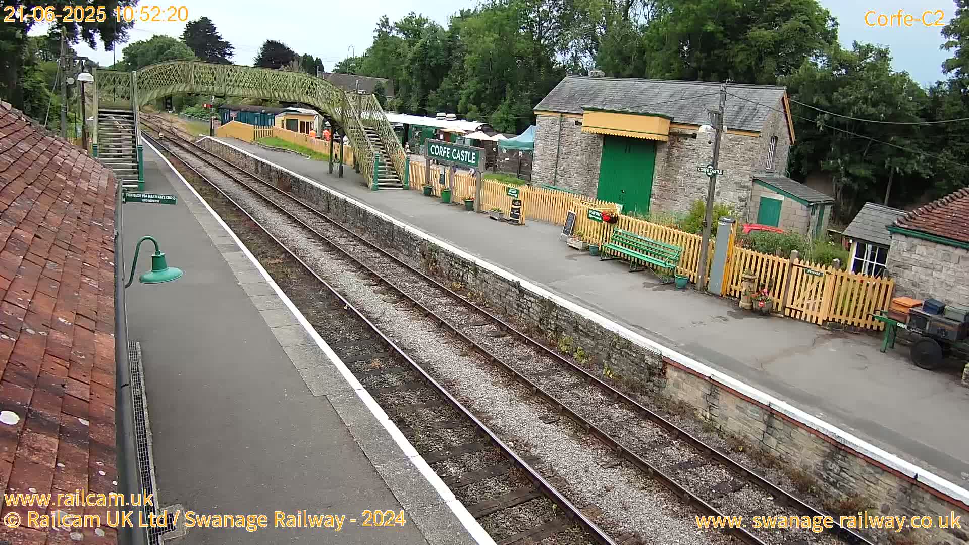 A railway station platform with two train tracks, a footbridge, and several stone buildings under a partly cloudy sky.