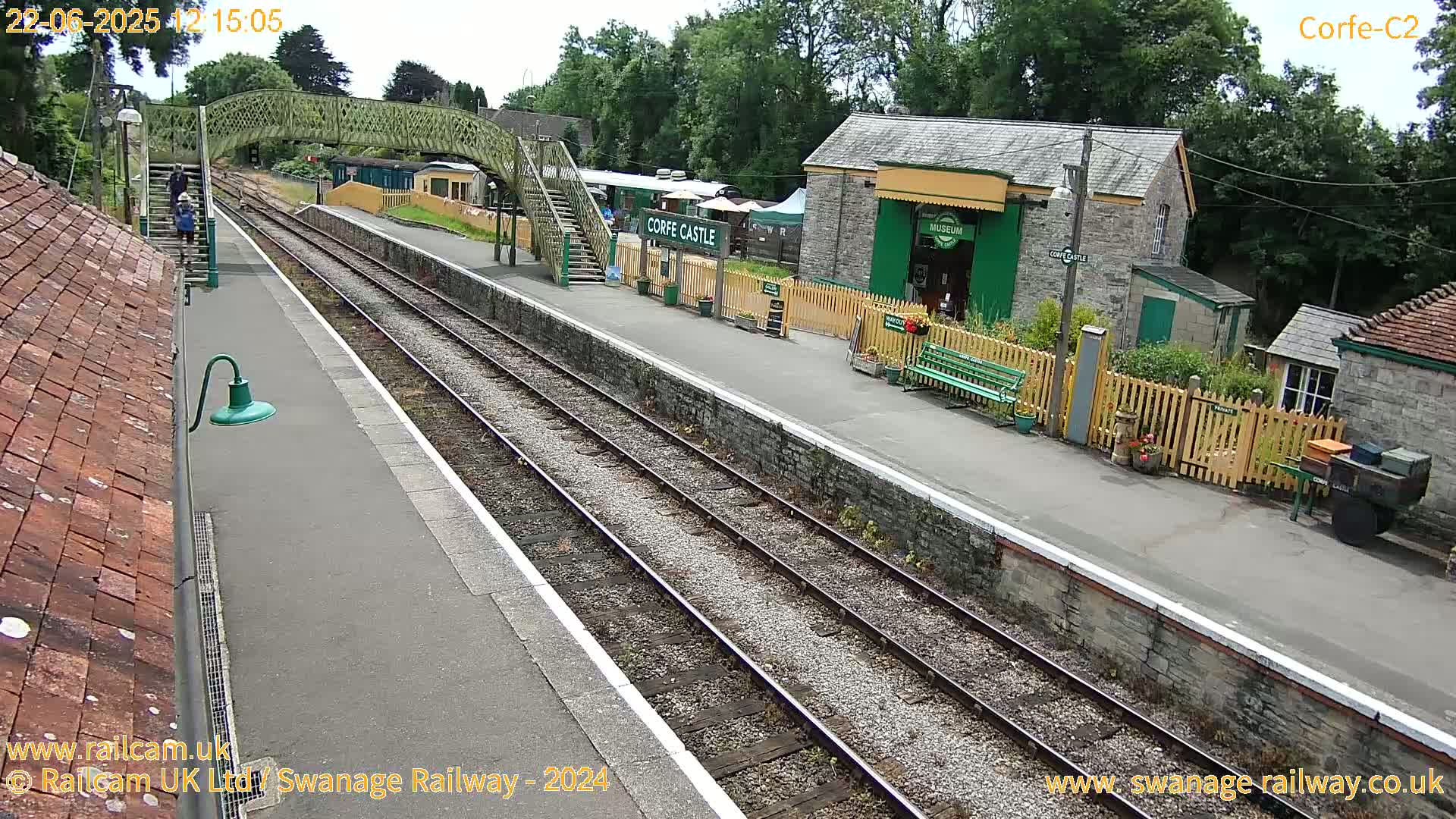 A train station platform with two sets of tracks, a footbridge, and several small buildings under a partly sunny sky.