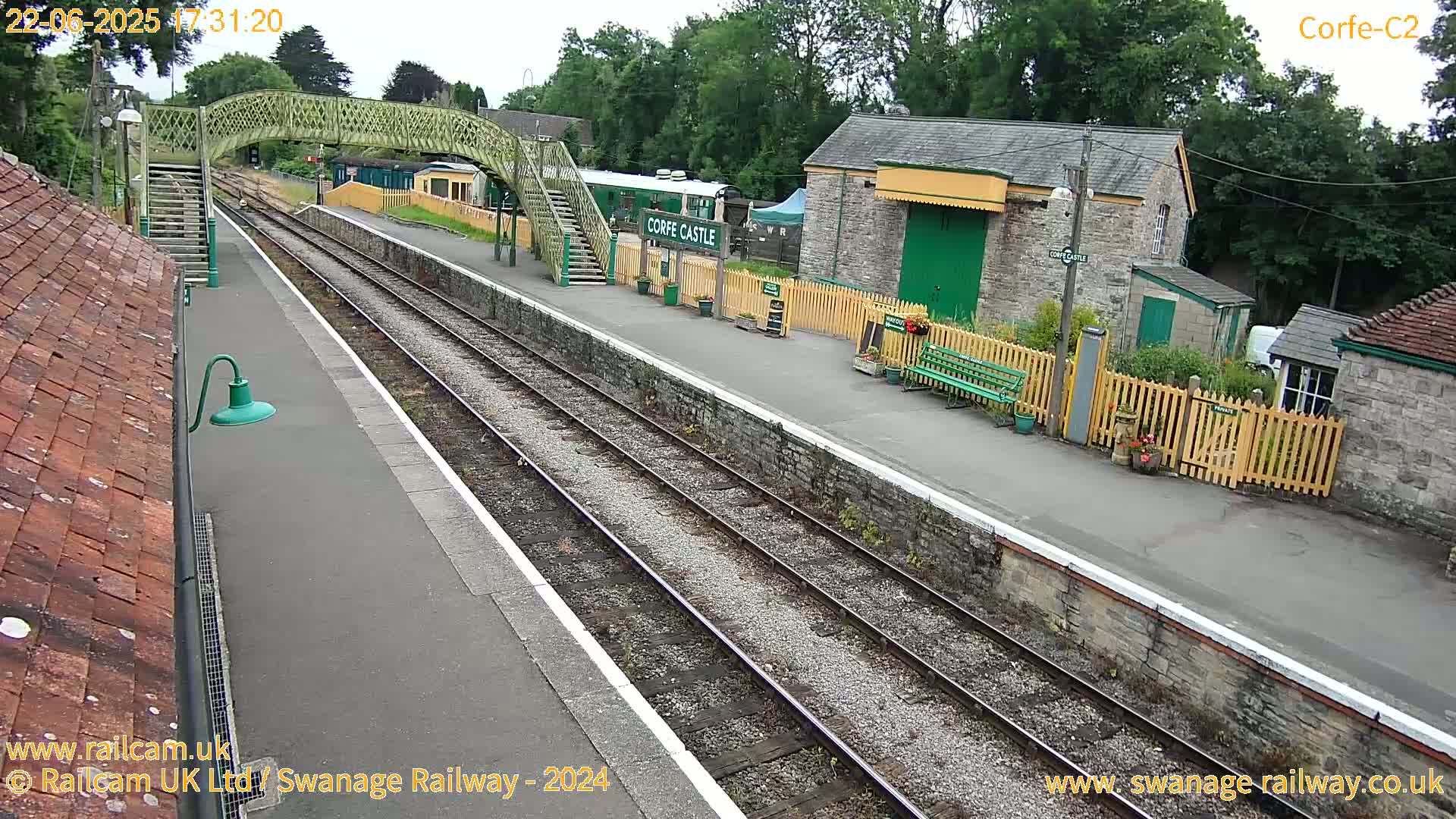 A train station platform with railway tracks, a footbridge, and several stone buildings under an overcast sky.