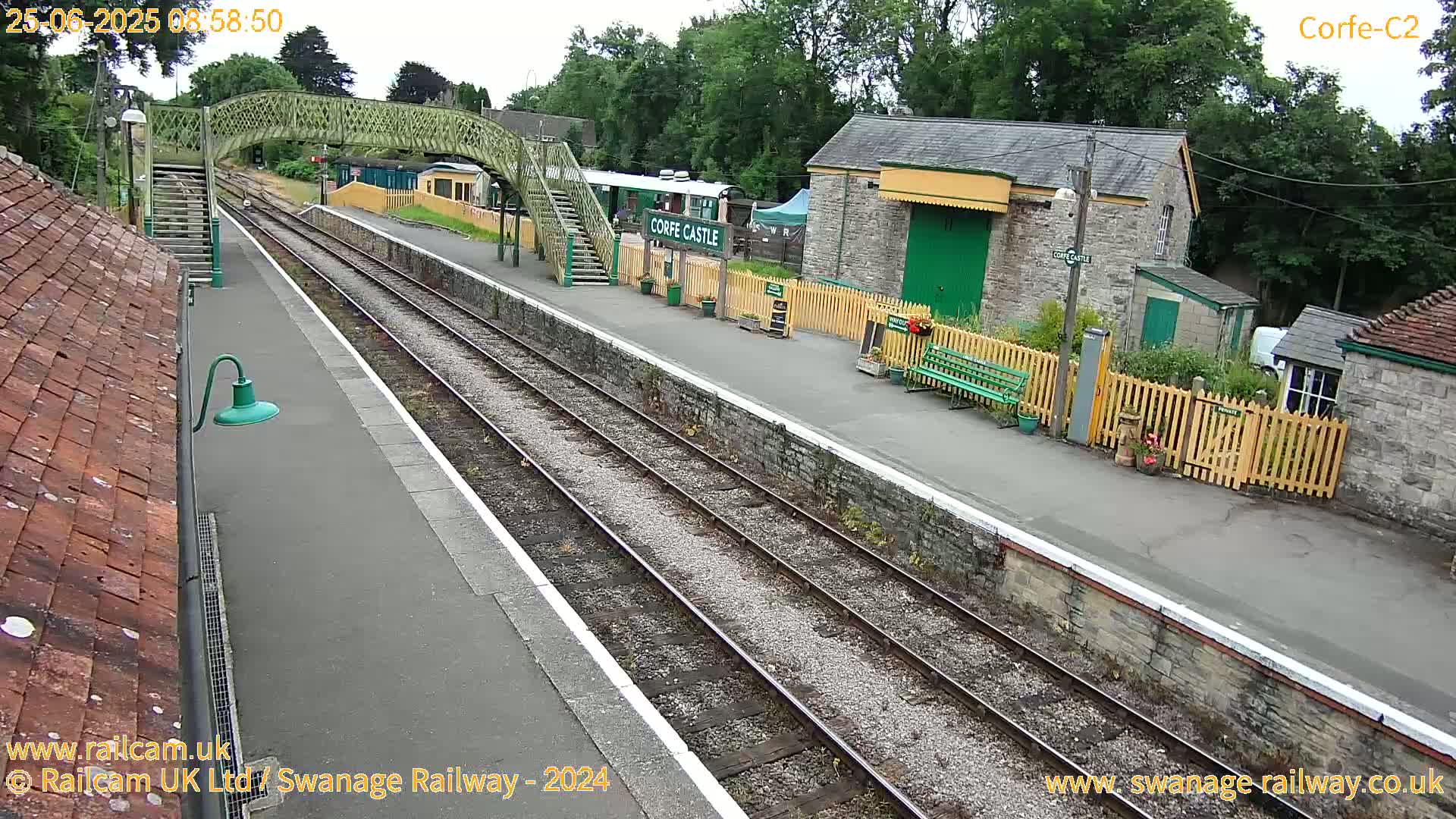 A train station platform with a footbridge, stone buildings, and train tracks under an overcast sky.