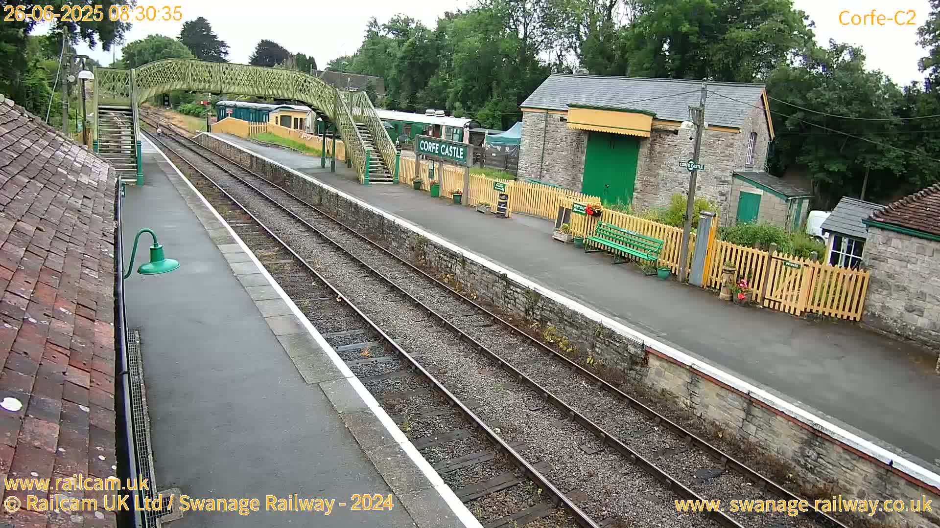 A train station platform with two train tracks, a pedestrian bridge, and several stone buildings is visible on an overcast day.