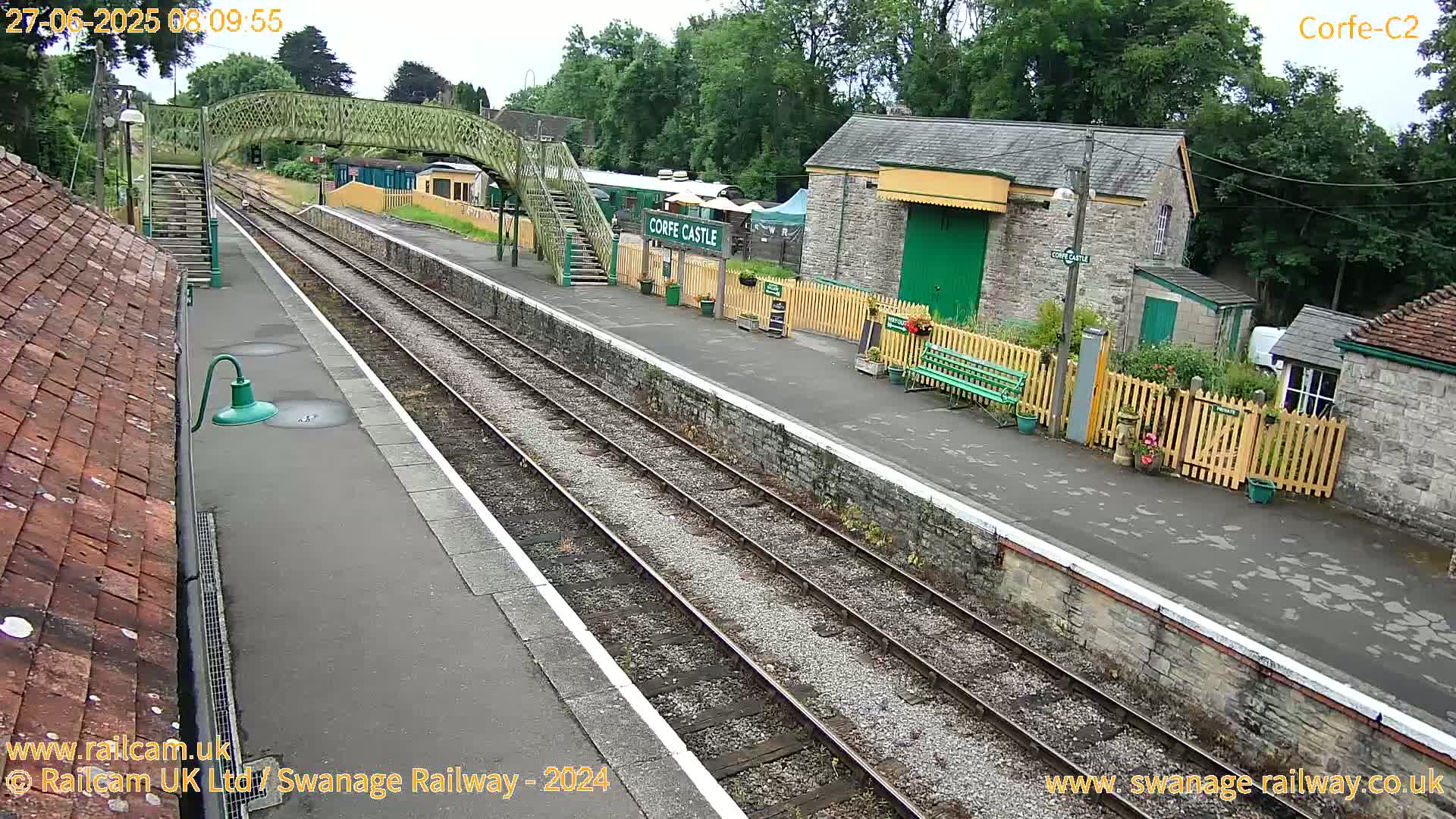 A train station platform with two train tracks, a footbridge, and several stone buildings is visible on an overcast day.