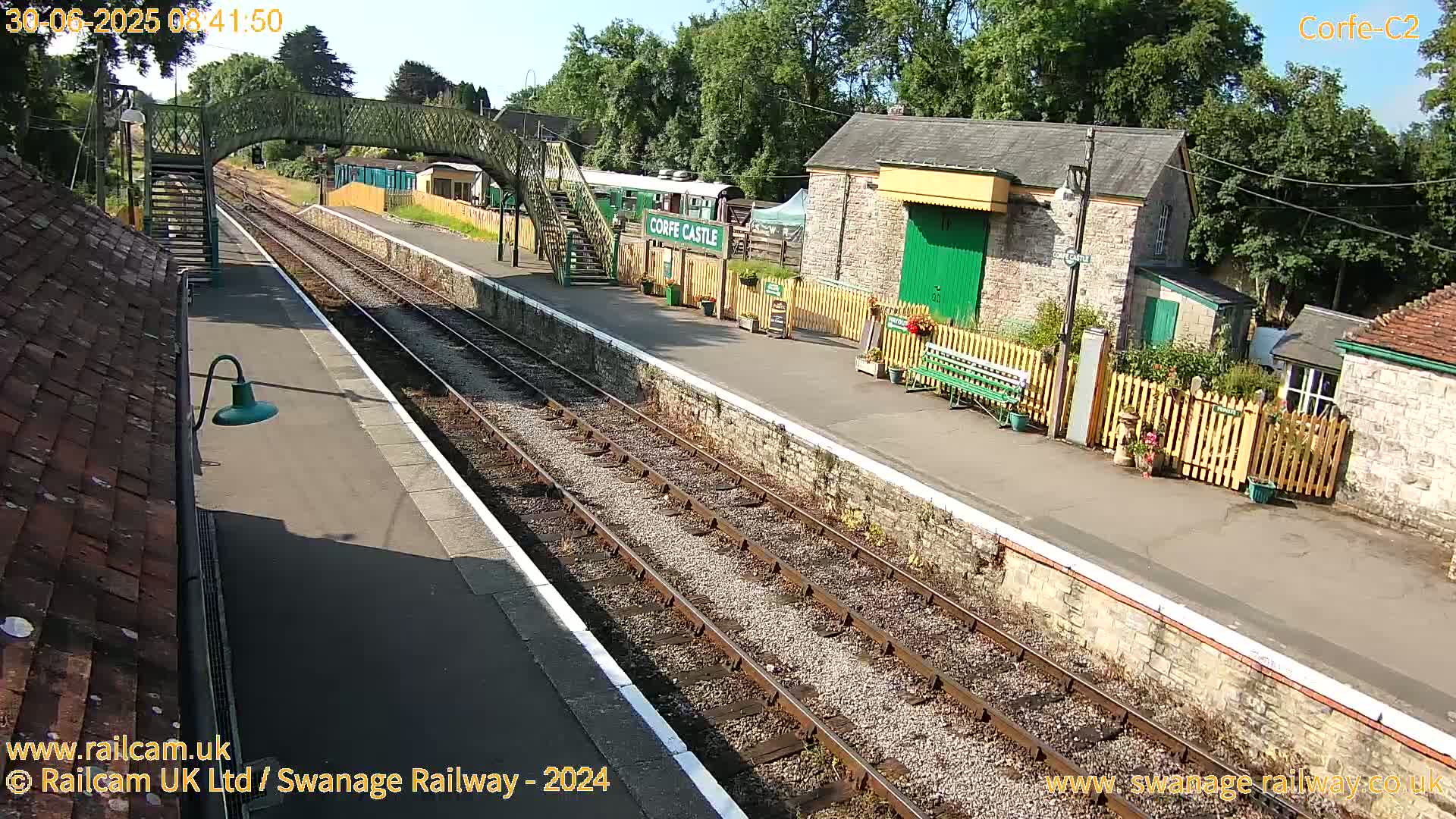 A sunny view of a train station platform with railway tracks, stone buildings, and a footbridge.