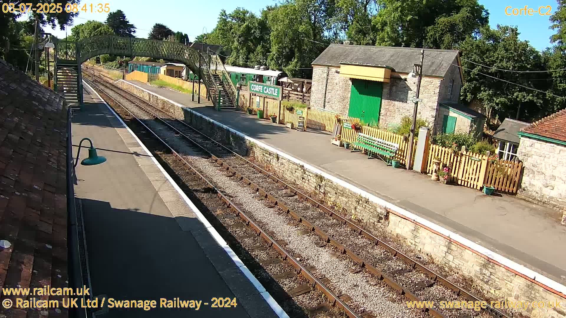 A sunny view of a train station with multiple tracks, a stone building, and a pedestrian bridge.