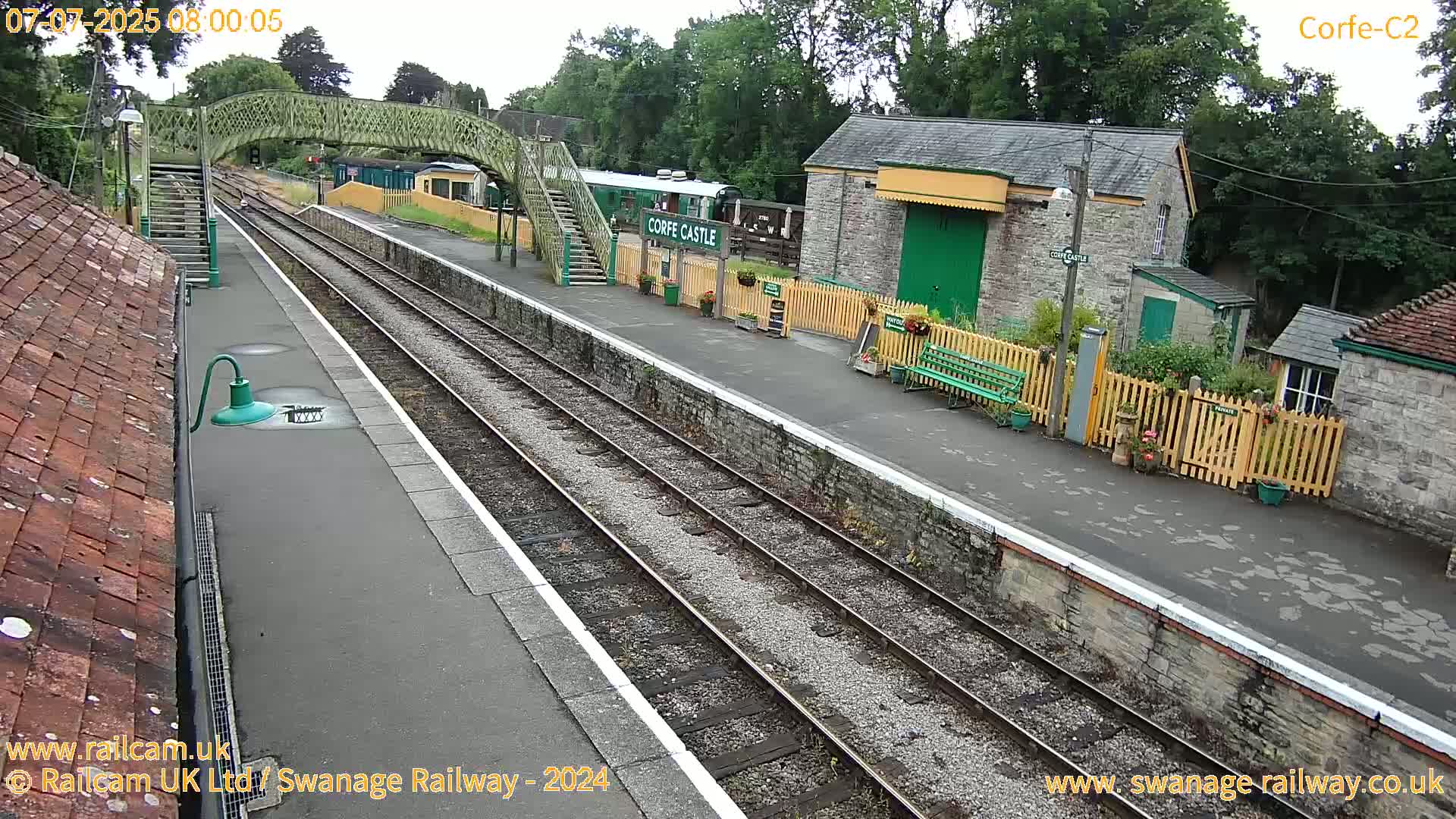 A train station platform with multiple train tracks under an overcast sky, featuring a pedestrian bridge, a railway carriage, and stone buildings.