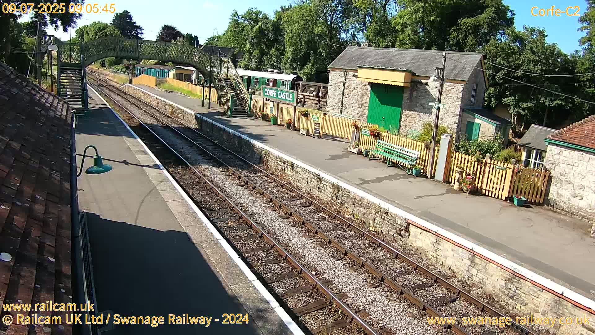 A sunny view of a train station with railway tracks, stone buildings, a footbridge, and a railway carriage.