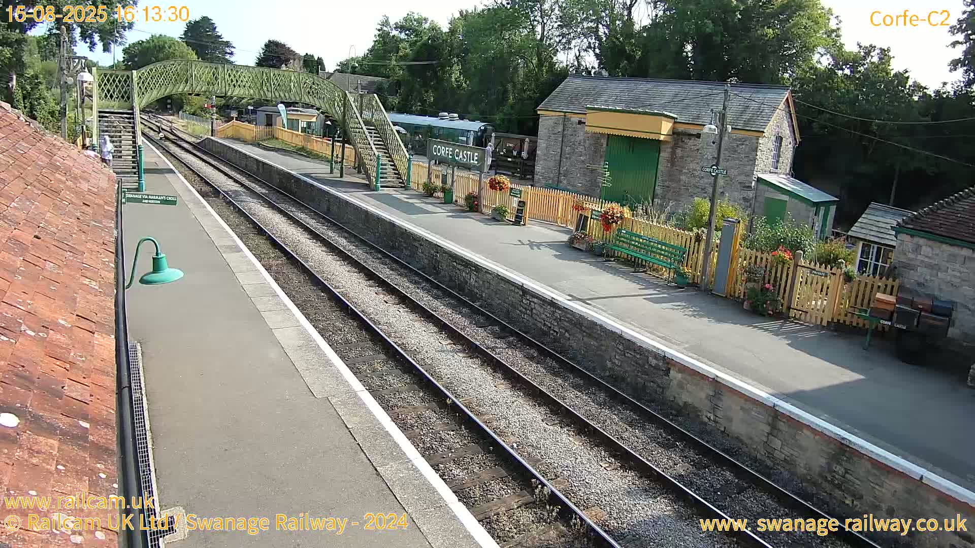 A sunny view of a train station with railway tracks, stone buildings, a footbridge, and a railway carriage.