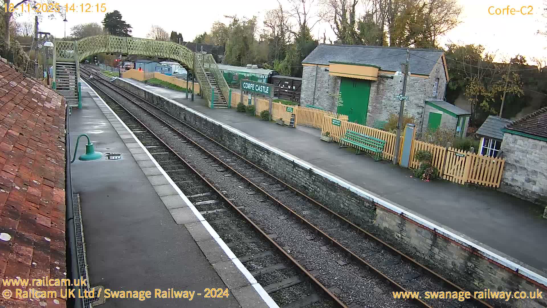 A sunny view of a train station with railway tracks, stone buildings, a footbridge, and a railway carriage.