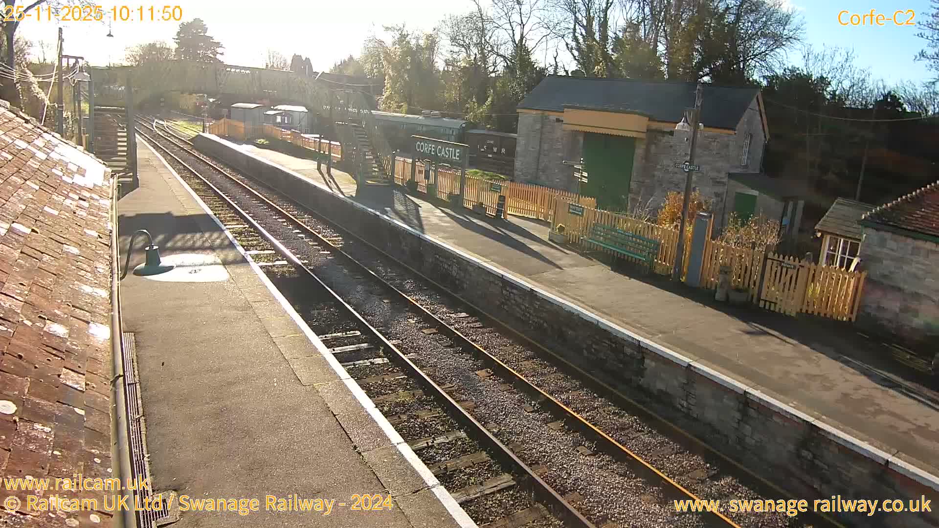 The image depicts a sun-drenched train station with empty platforms and multiple tracks, featuring a green train carriage labeled "Corfe Castle" at the far platform, surrounded by various station buildings and mostly bare trees under a clear, sunny sky.