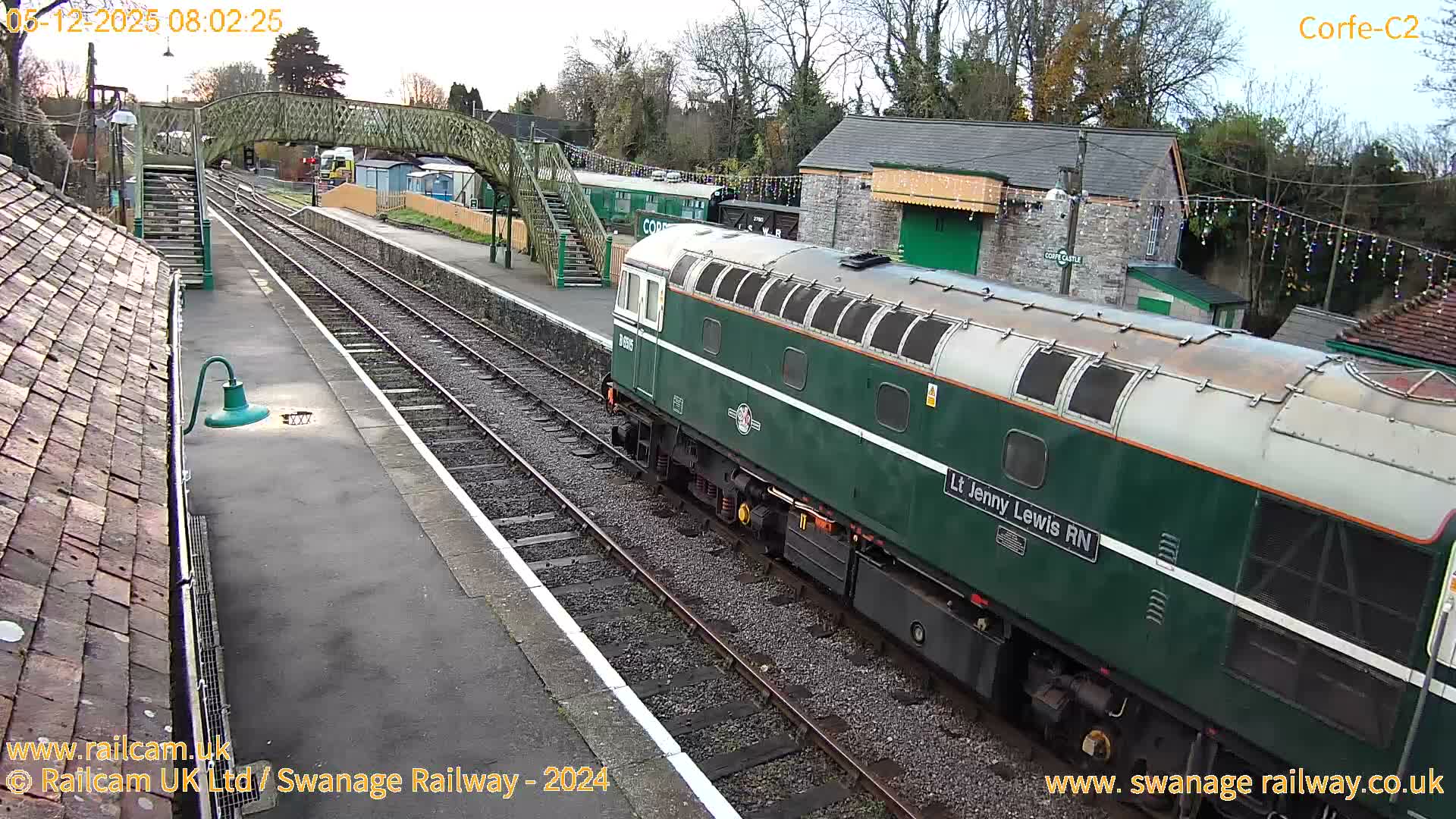 A dark green and cream locomotive is seen on tracks at an outdoor train station with a footbridge, platforms, and buildings decorated with string lights, under a bright, overcast sky.