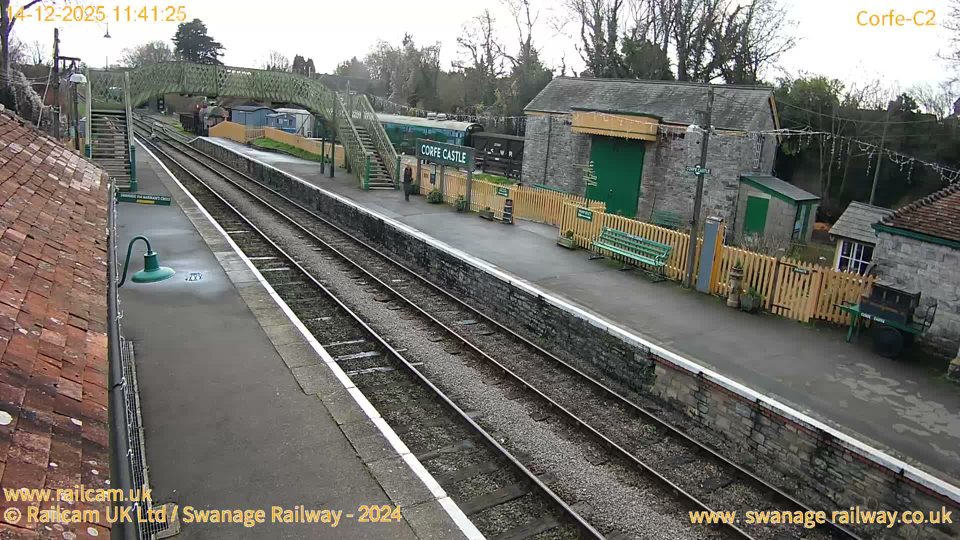 A dark green and cream locomotive is seen on tracks at an outdoor train station with a footbridge, platforms, and buildings decorated with string lights, under a bright, overcast sky.