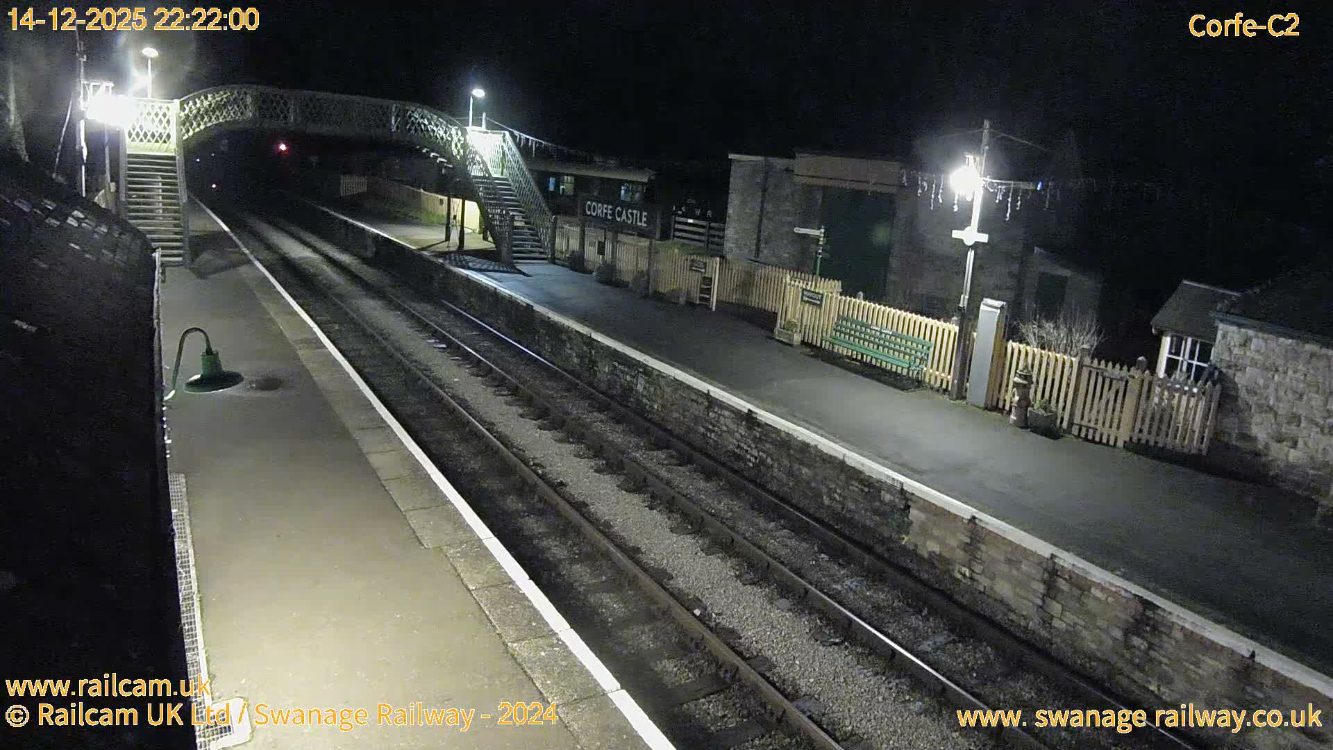 A dark green and cream locomotive is seen on tracks at an outdoor train station with a footbridge, platforms, and buildings decorated with string lights, under a bright, overcast sky.