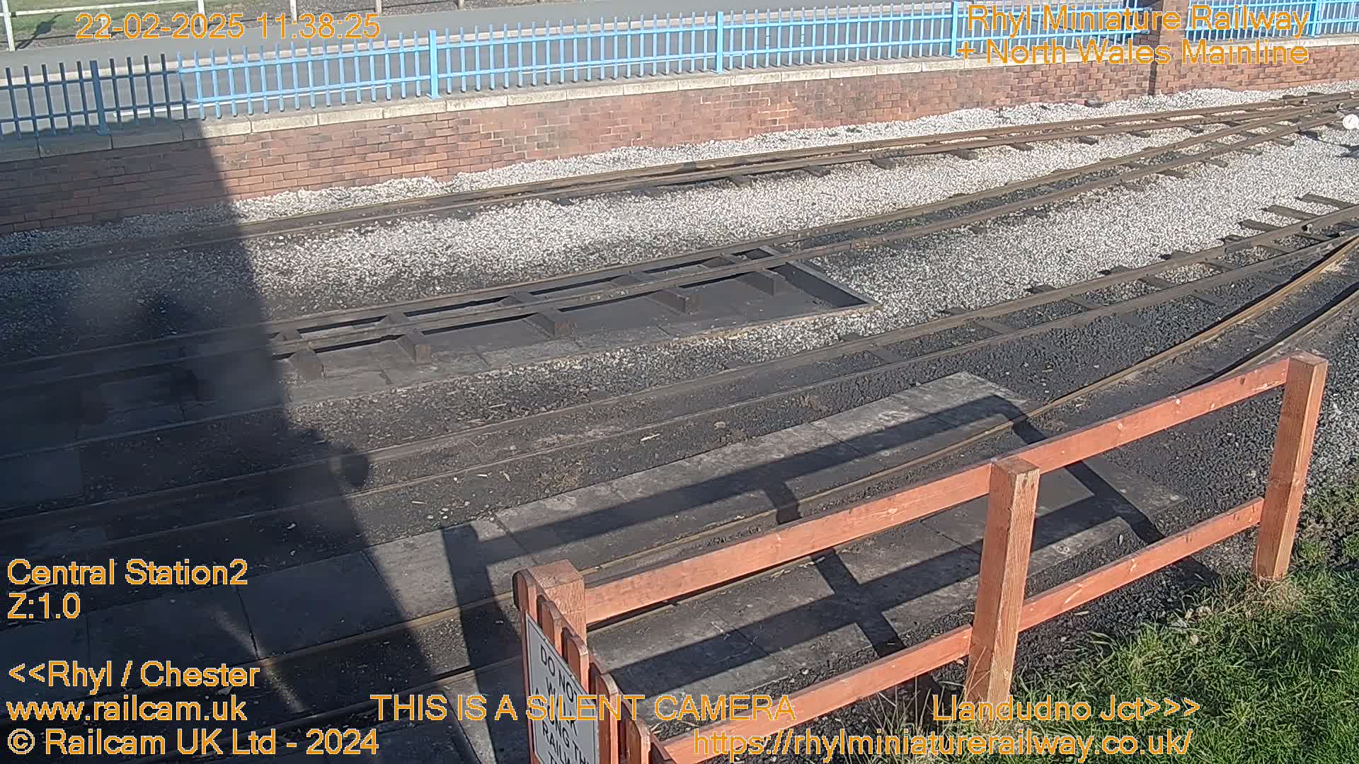 A sunny outdoor view of railway tracks and a wooden fence, with a long shadow cast across the scene.