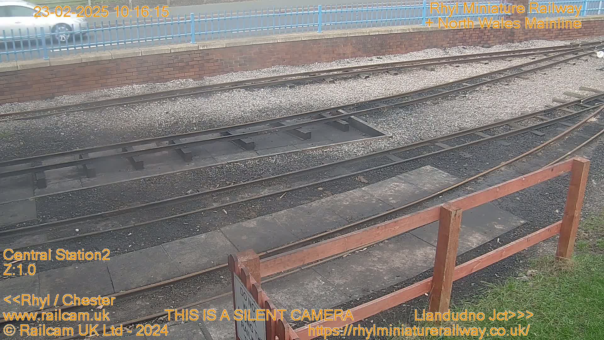 A high-angle view of a railway switching yard with several sets of tracks, gravel ballast, and a short wooden fence, under overcast skies.