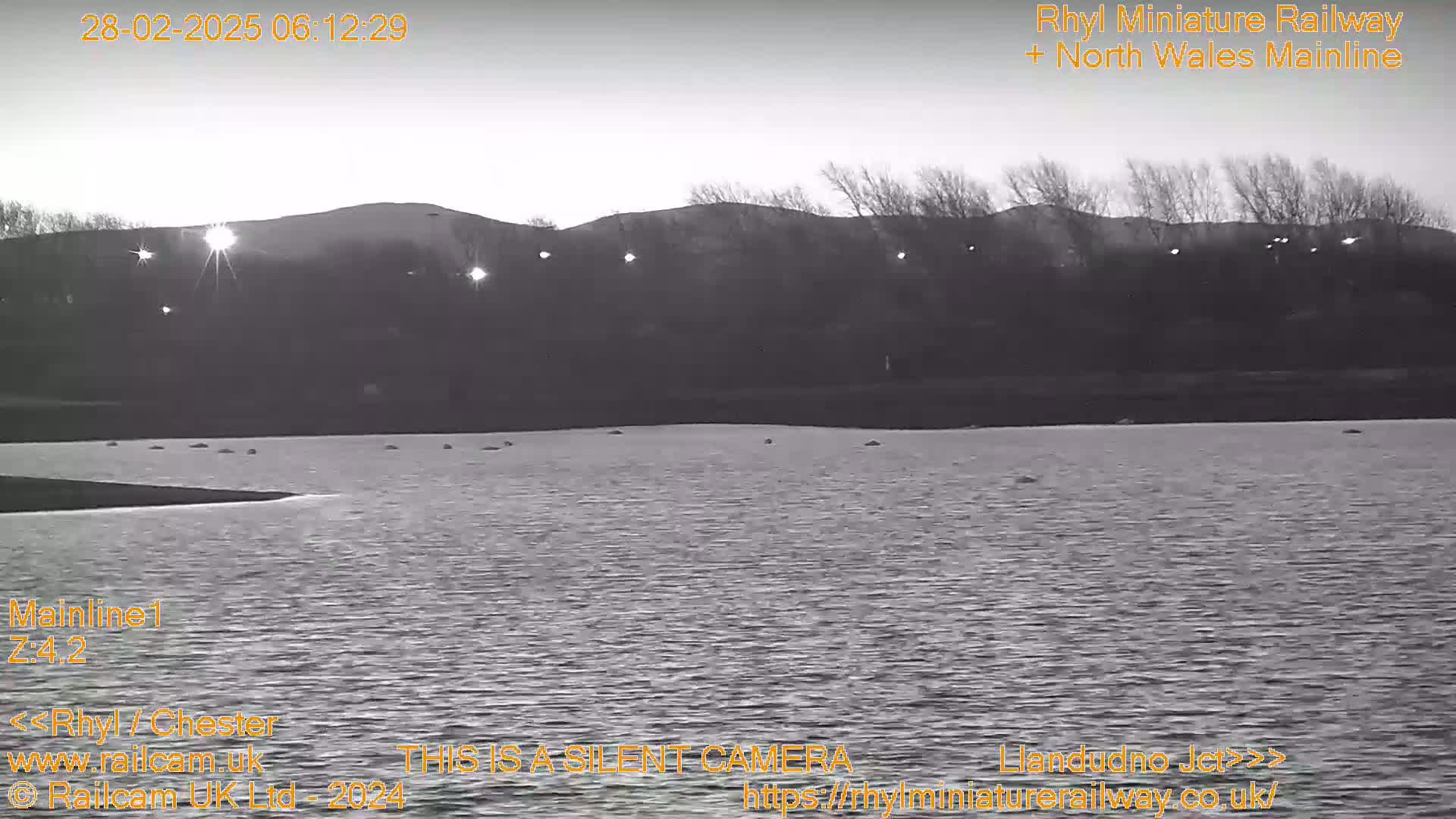 A grayscale image shows a calm body of water under a sunrise sky with distant hills and bare trees in the background.