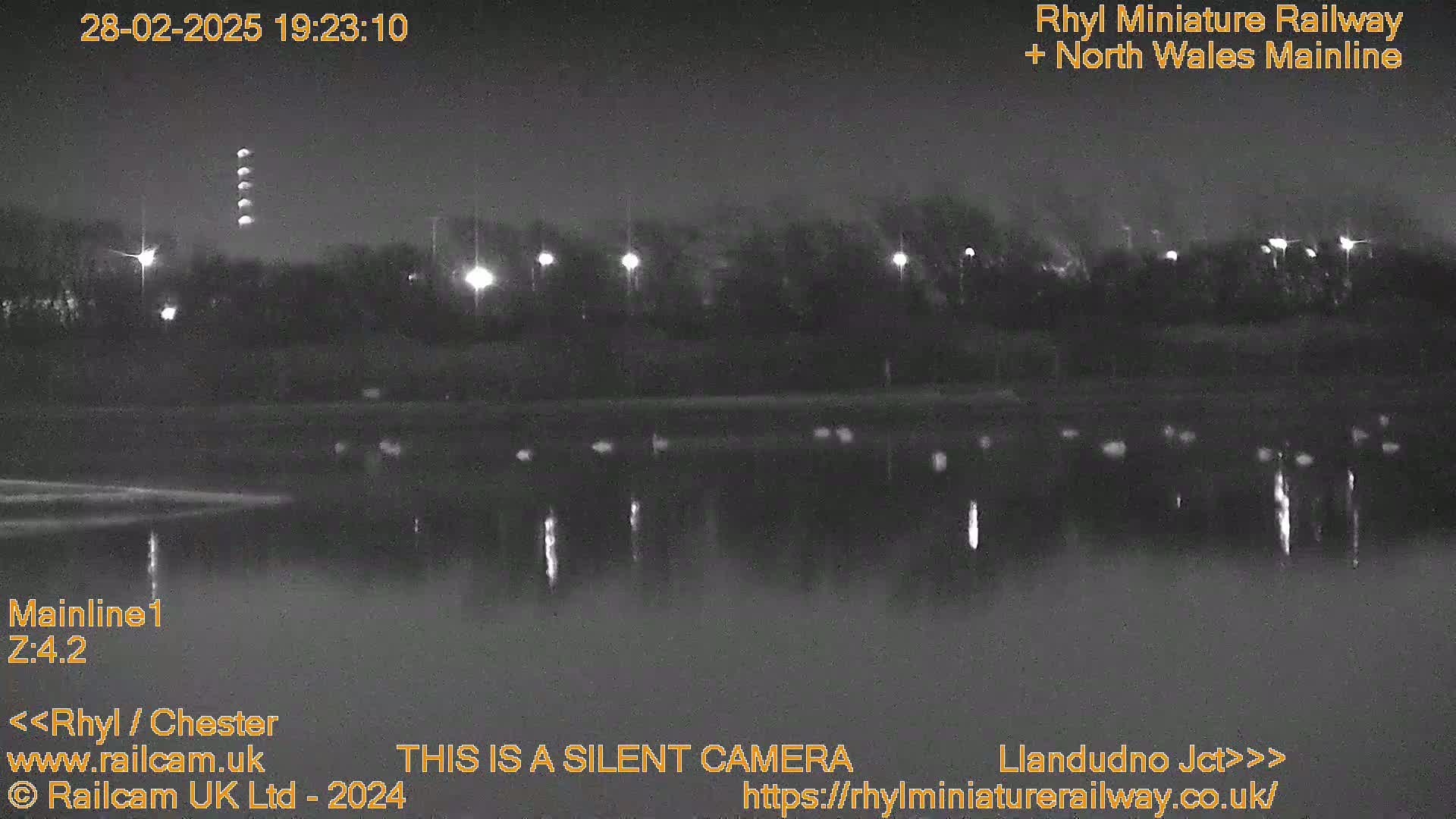 A nighttime, low-light view of a dark body of water reflecting lights from a distant shoreline with trees and lights.