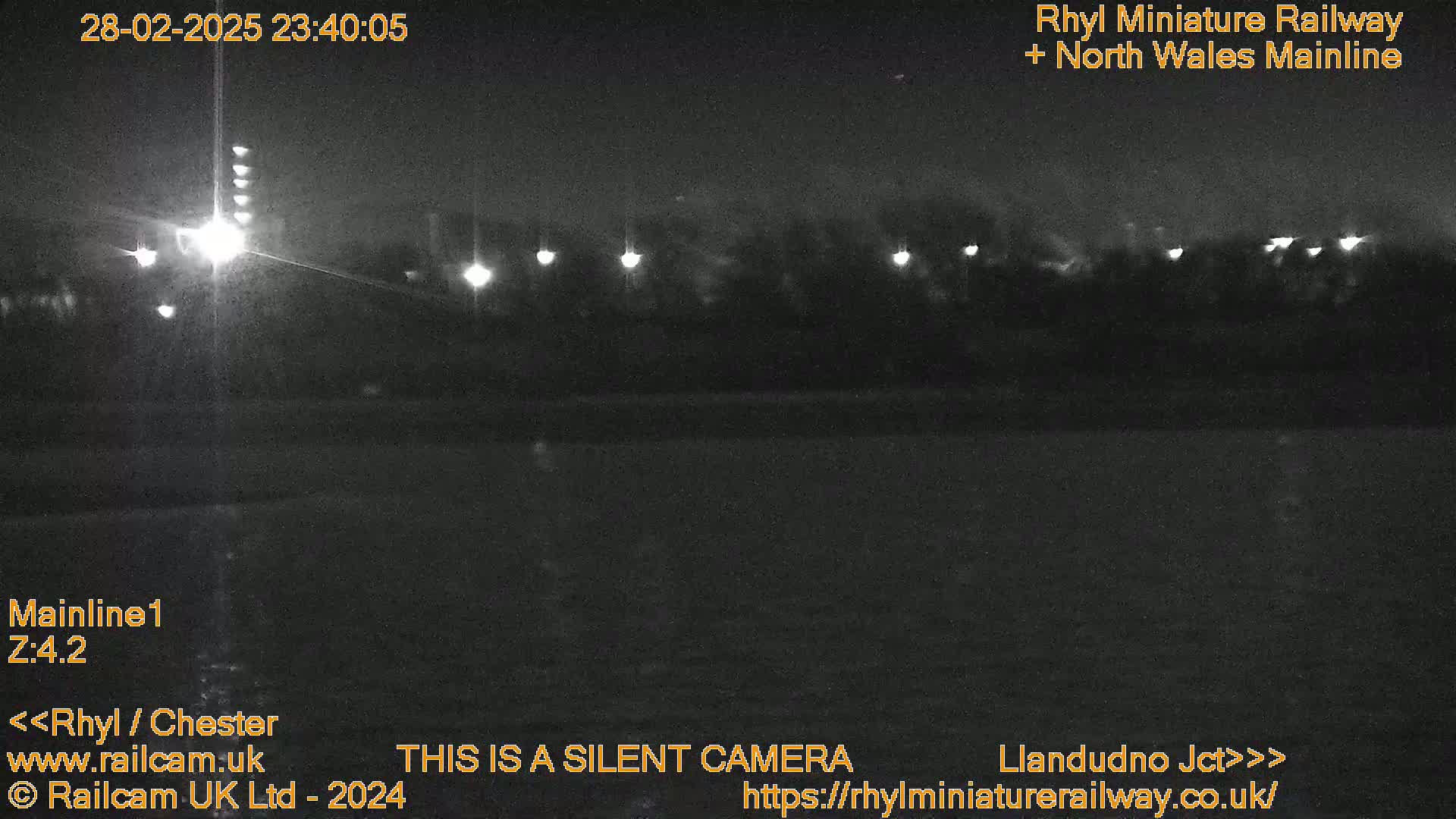 A nighttime, low-light view of a dark landscape with several distant lights illuminating trees and a flat area.