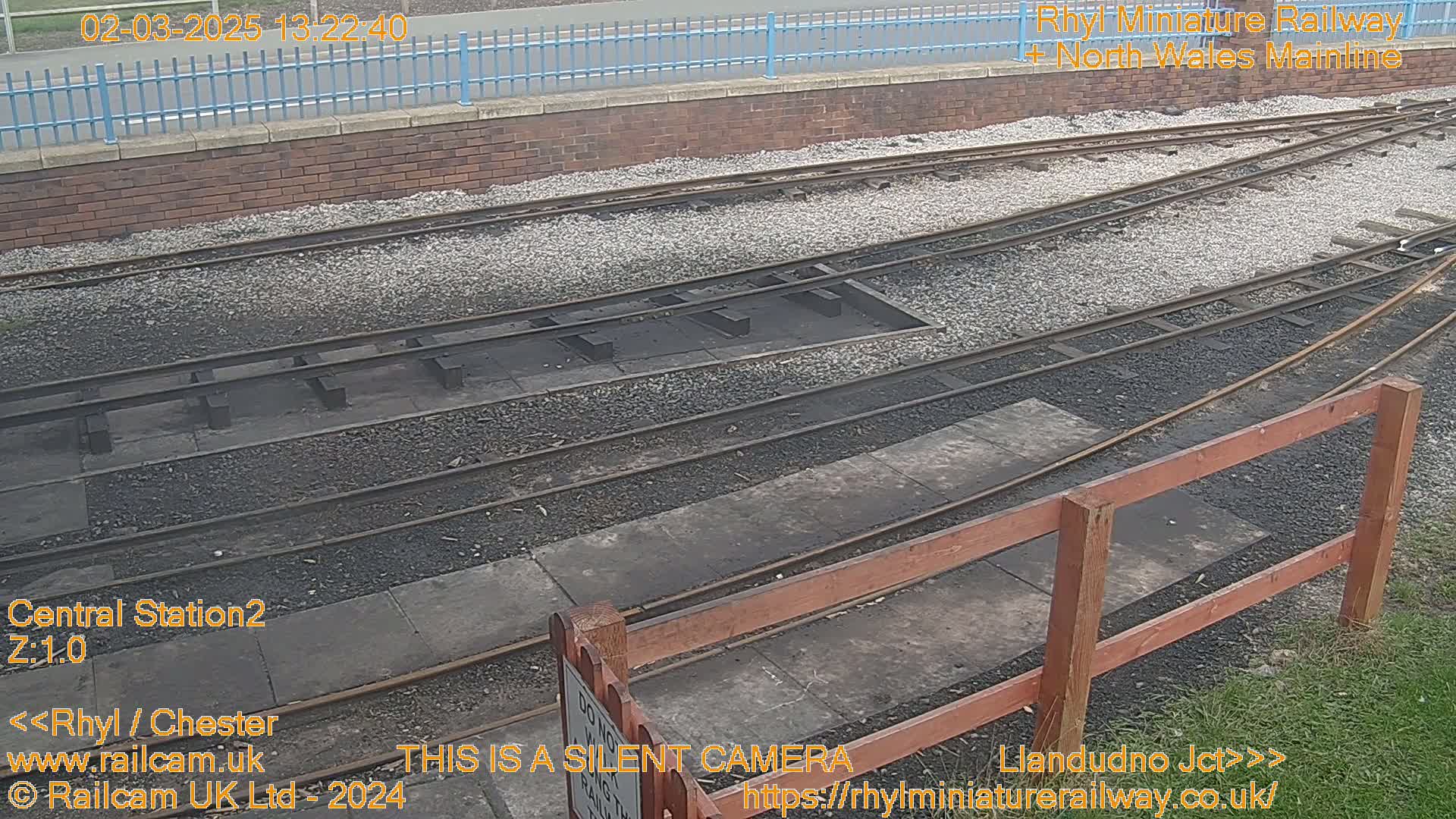 A section of railway tracks with a wooden fence and a brick wall in the background under an overcast sky.