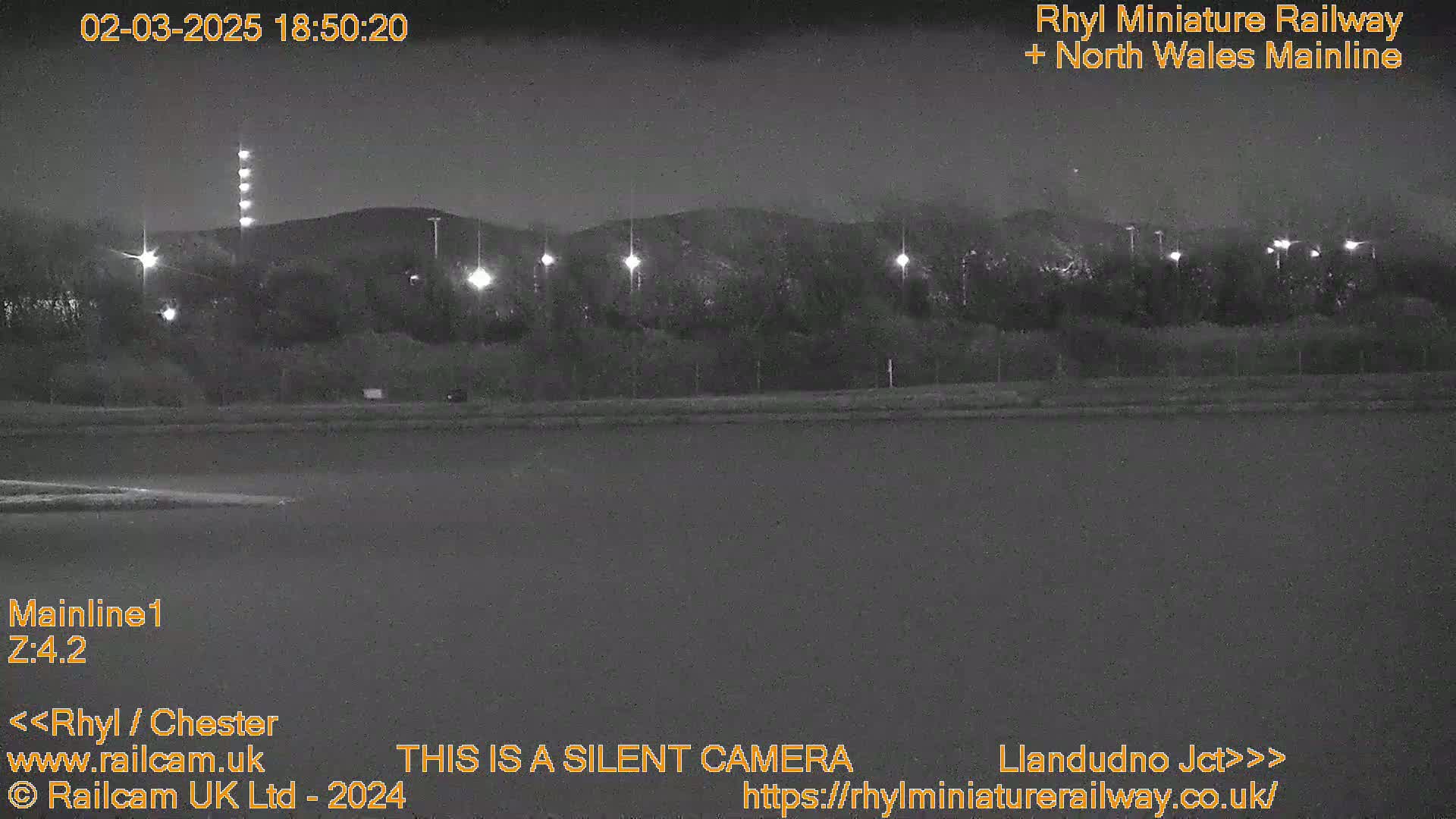 A nighttime, low-light view of a dark body of water, a roadway with illuminated lights, and a treeline silhouetted against a hill in the background.