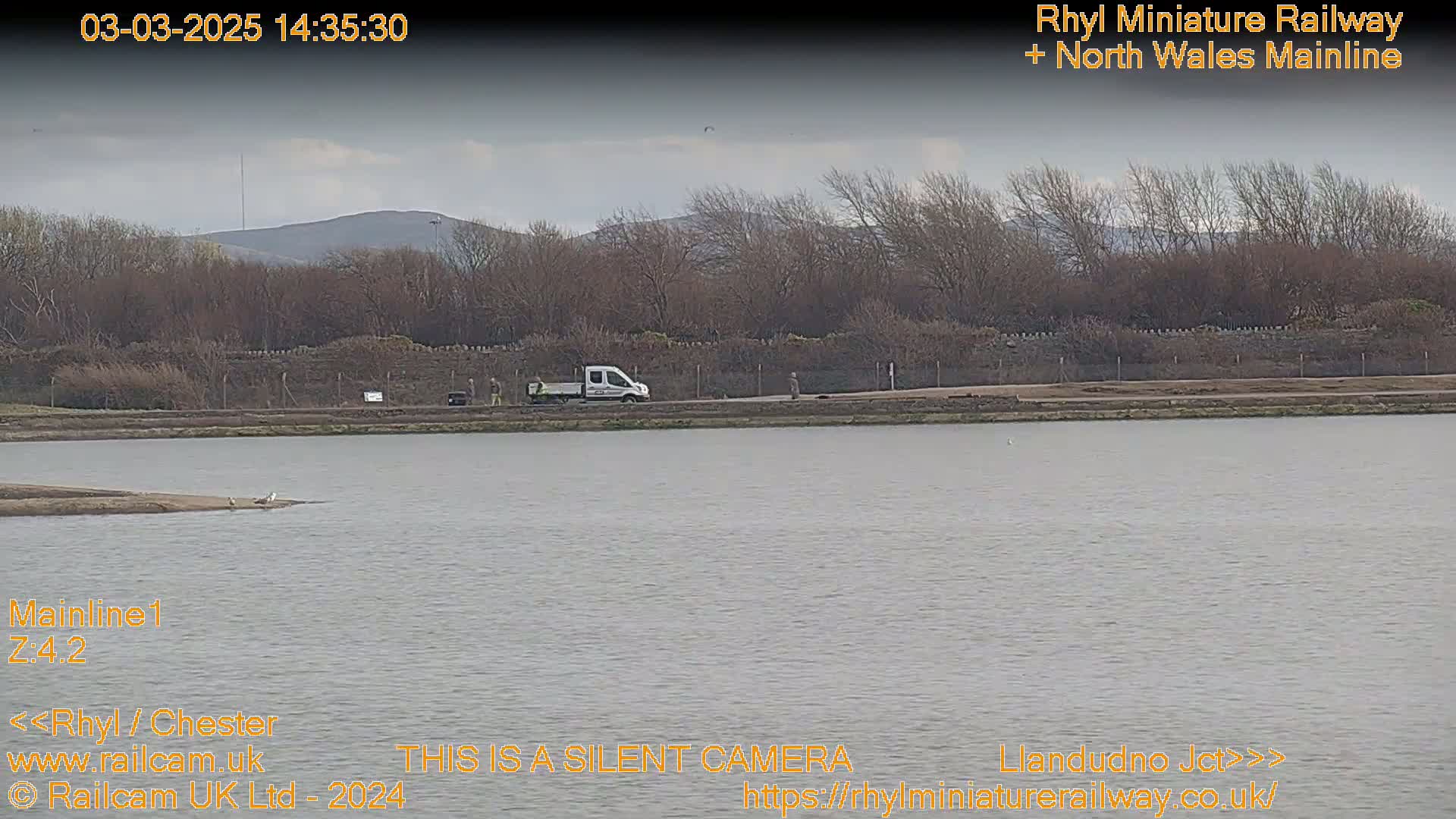 A white van is parked near a body of water under a partly cloudy sky, with bare trees and distant hills in the background.