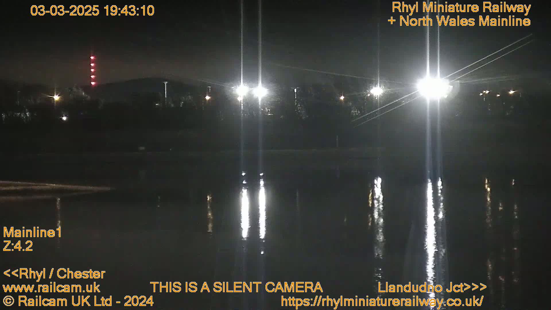 At night, bright lights illuminate a dark body of water reflecting the lights, with dark trees and a tall structure with red lights visible in the distance.