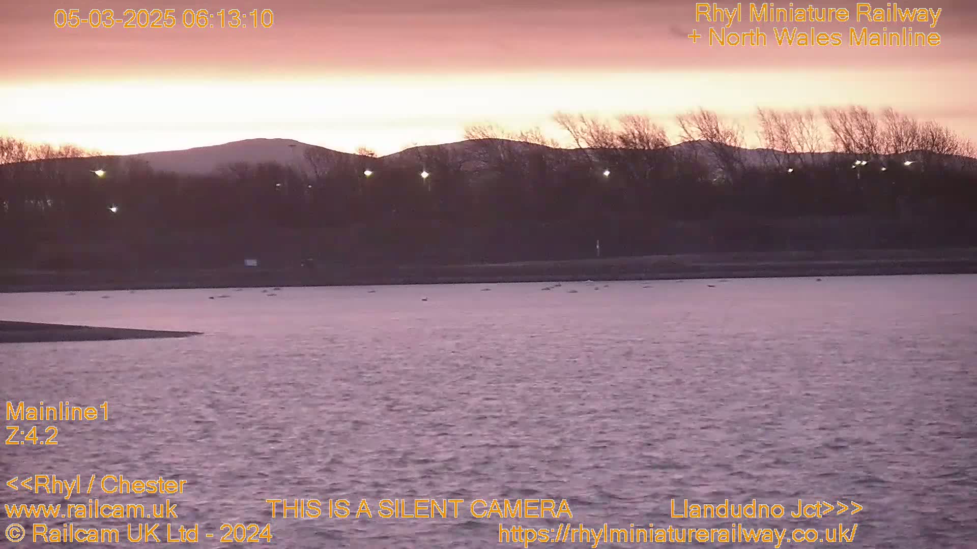 A calm body of water sits before a line of trees and hills under a pinkish sunrise.