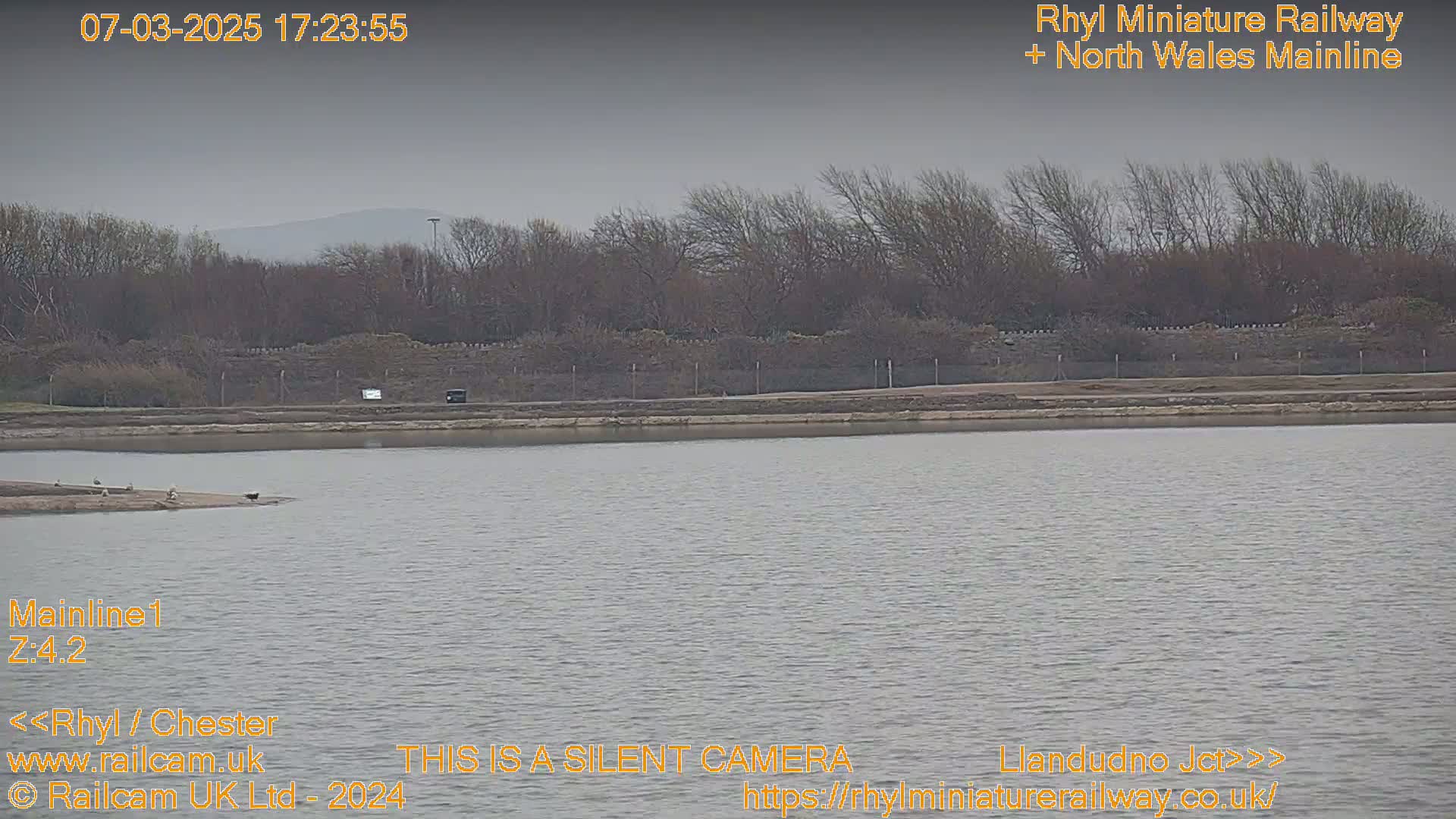 A body of still water stretches out before a line of bare trees and a low stone wall under an overcast sky.