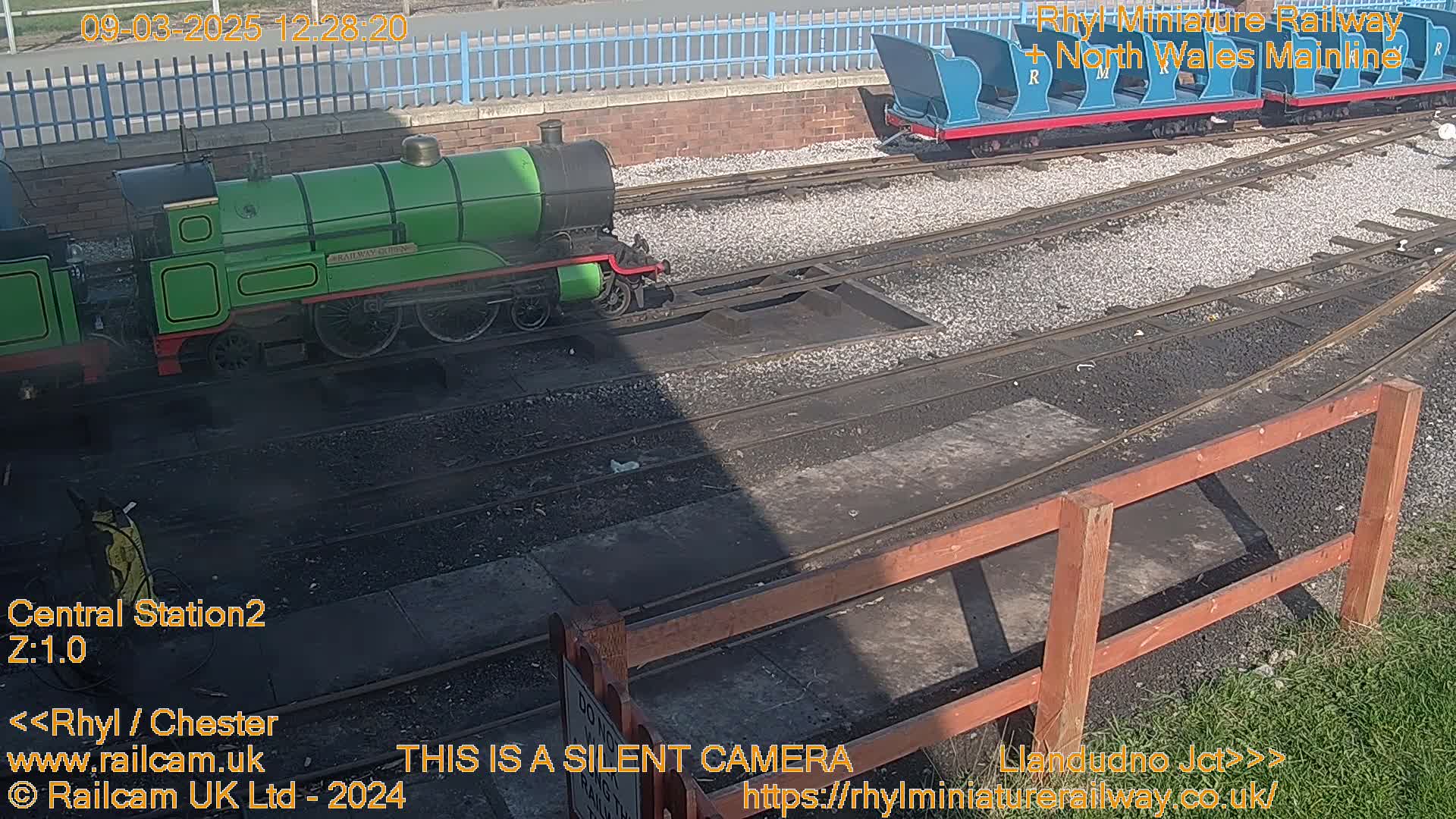 A green miniature train sits on a railway track next to a section of blue railway cars, all under a sunny sky.