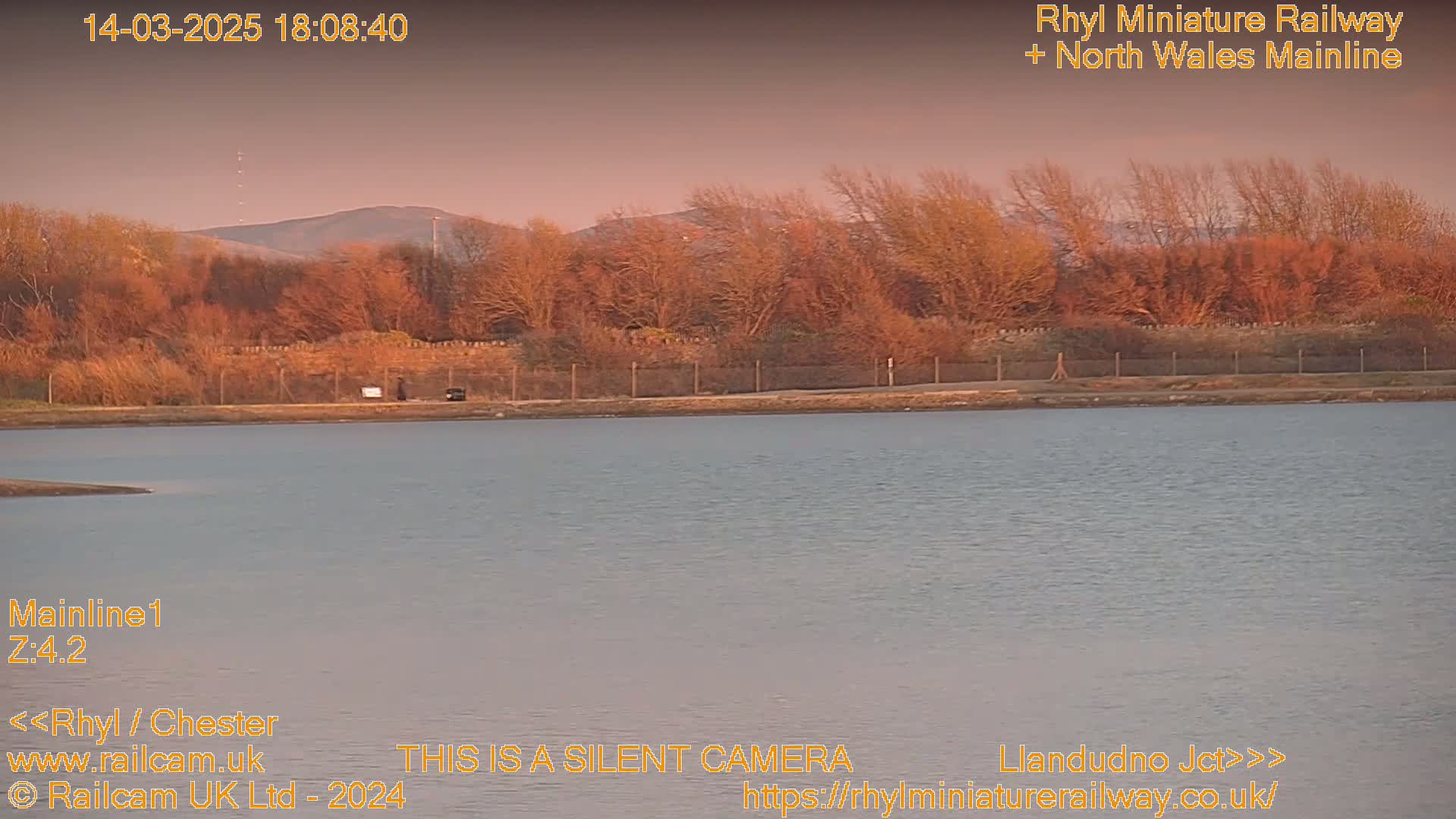 A calm body of water sits before a line of bare, reddish-brown trees silhouetted against a dusky pink sky and distant hills under a clear evening sky.
