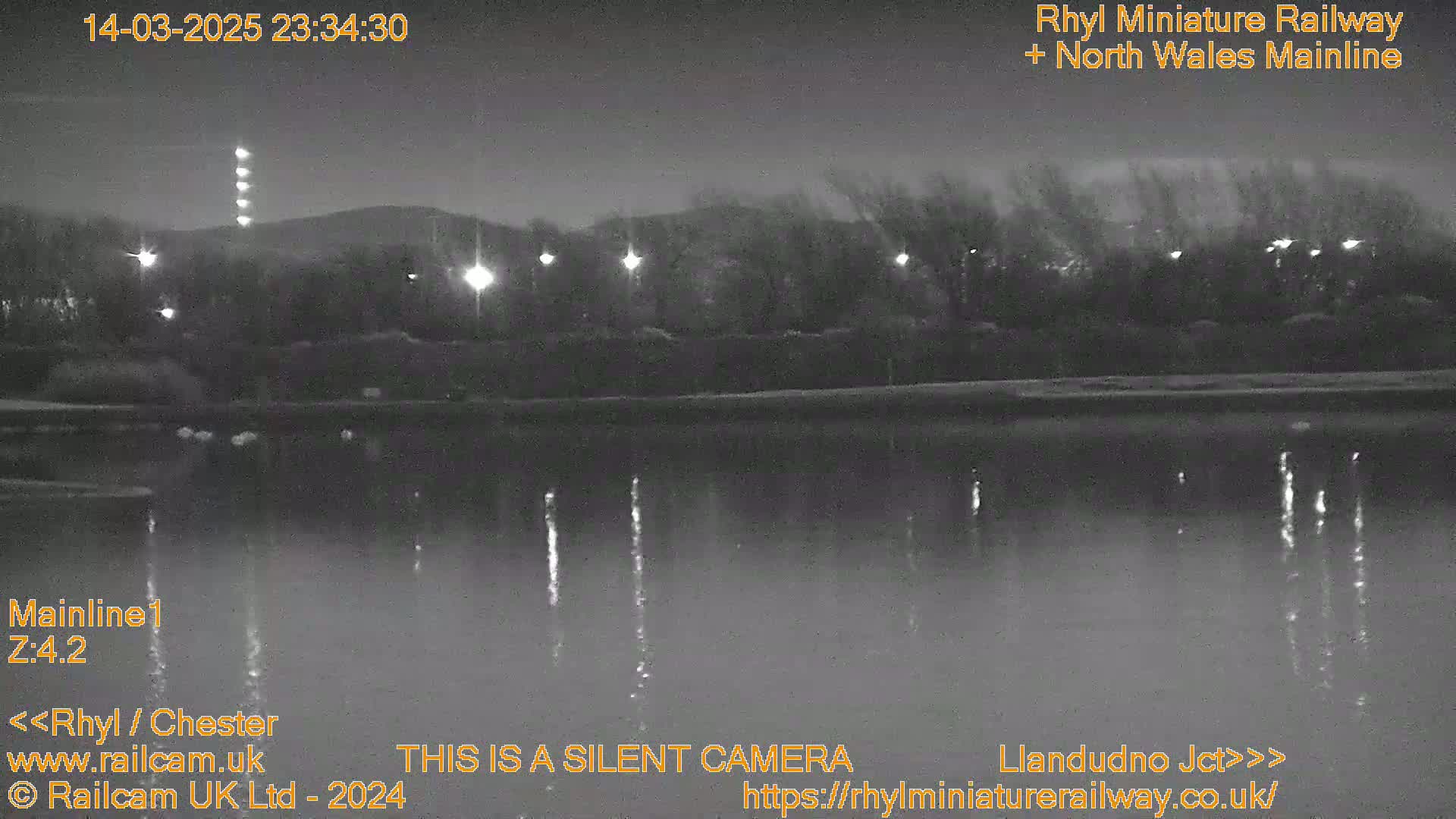 A nighttime, low-light view of a calm body of water reflecting lights from the opposite shore, with a dark landmass and trees in the background.