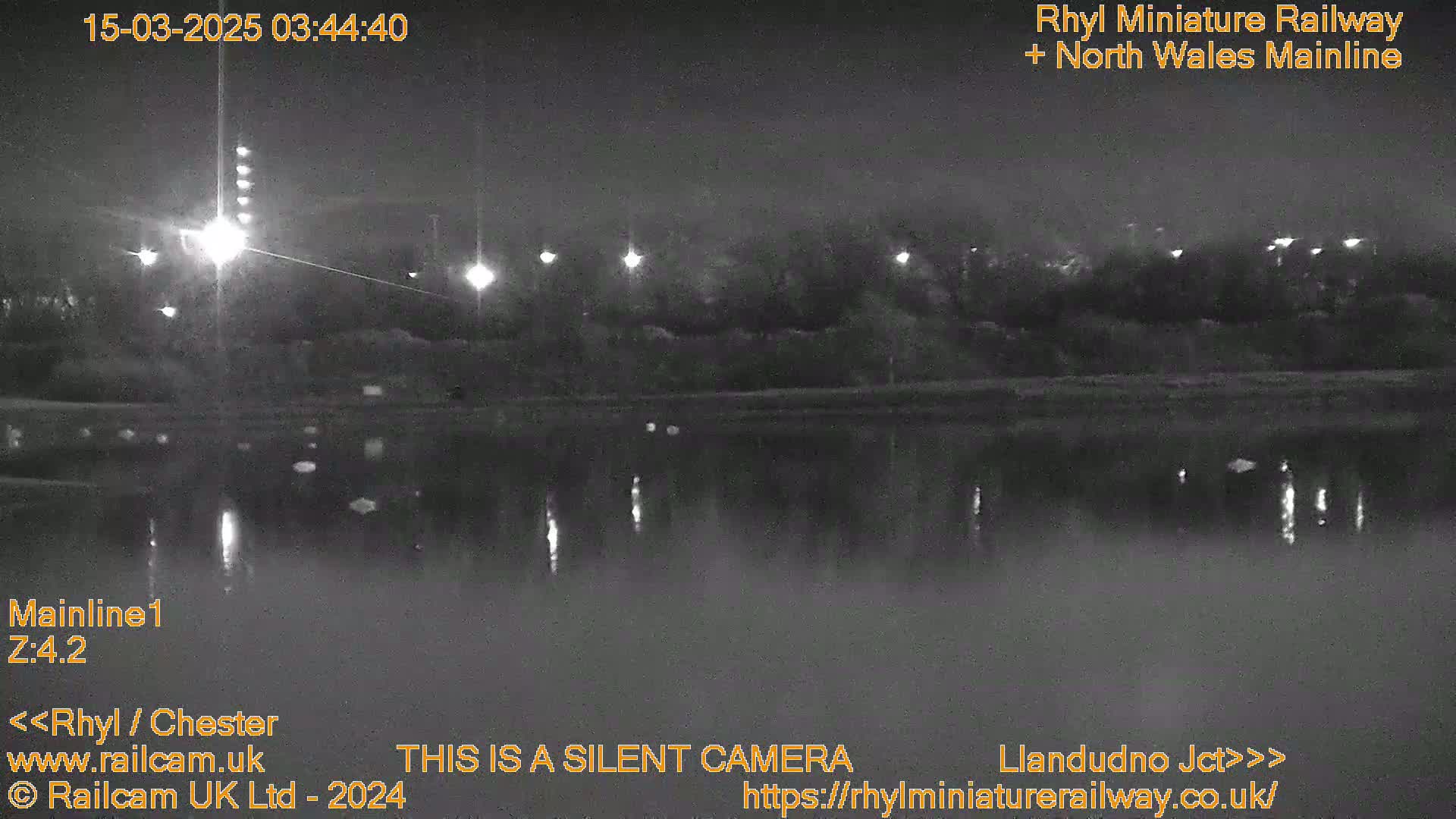 A nighttime, low-light view of a body of calm water reflecting lights from the opposite bank, with dark trees and foliage visible on the far shore.