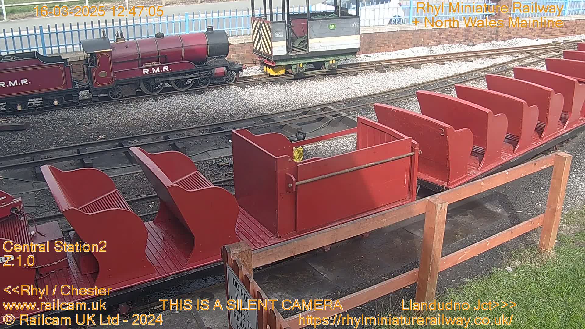 A red miniature train, consisting of an engine and several open-top passenger cars, sits on a track outdoors on a sunny day.