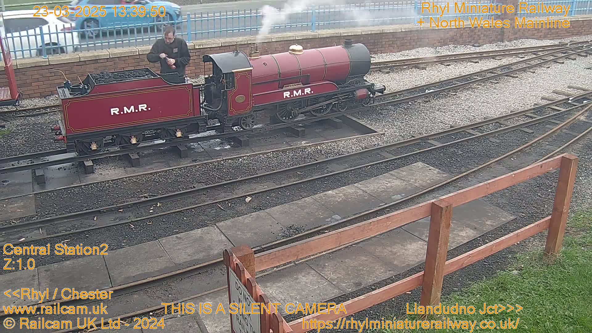 A red miniature steam train is moving along a track, with a person tending to it, in what appears to be an outdoor setting.