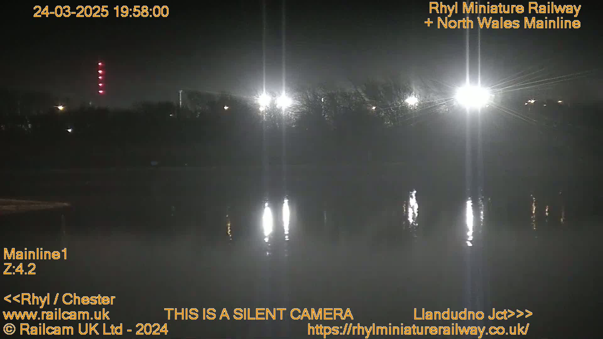 At night, bright lights illuminate a dark body of water reflecting the lights, with trees and other vegetation visible along the water's edge.